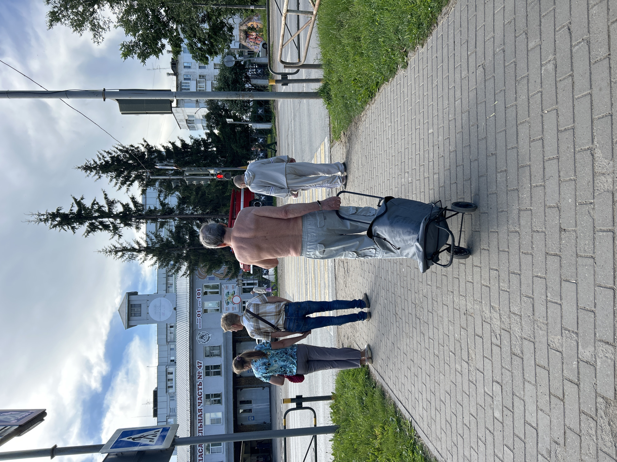 In Chaikovsky, Russia, on a paved sidewalk under a partly cloudy sky, a shirtless man with gray hair walks away from the camera, pulling a wheeled, dark gray fabric bag. He is wearing loose-fitting, light-colored cargo pants. To his left, a couple walks away from the camera, holding hands. The woman is wearing a blue and black patterned shirt and gray pants. The man is wearing a plaid shirt with a shoulder bag and blue jeans. Behind them, a building with a sign that reads "ПОЖАРНАЯ ЧАСТЬ № 47" (Fire Station No. 47) is visible, with trees and other buildings in the background. A traffic light is red, and a crosswalk is marked on the street.