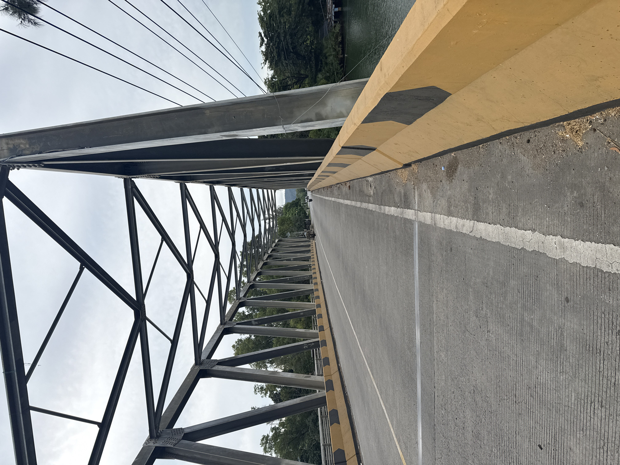 The image shows a view of a bridge spanning a body of water in Lamkruet, Indonesia. The bridge is constructed with a steel truss superstructure and a concrete deck. The road on the bridge has a yellow and black barrier on the right side, with a white lane divider. Trees and vegetation line the banks of the water. The sky is overcast. On the bridge, there are a few motorcycles and a person on foot visible in the distance. The scene appears to be during daylight hours.