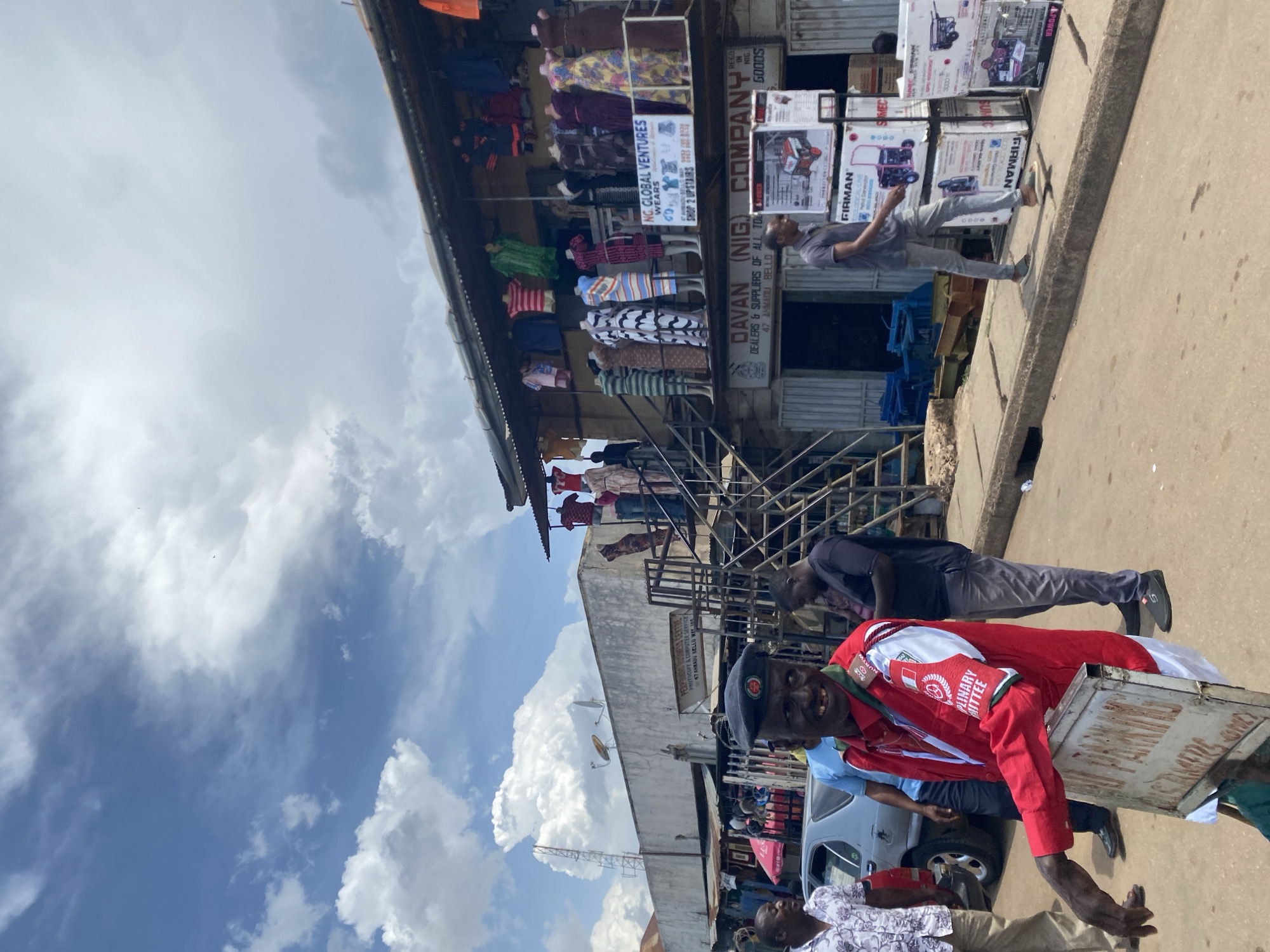 A street scene in Jos, Nigeria, features a man in a red jacket and a beret, smiling at the camera. He is holding a sign that reads "NO PARKING". Behind him, several people are visible, some walking and others standing around a building with multiple shops. The building has clothing displayed on mannequins on the upper level. A car is parked on the left side of the image. The sky is partly cloudy with bright sunlight.