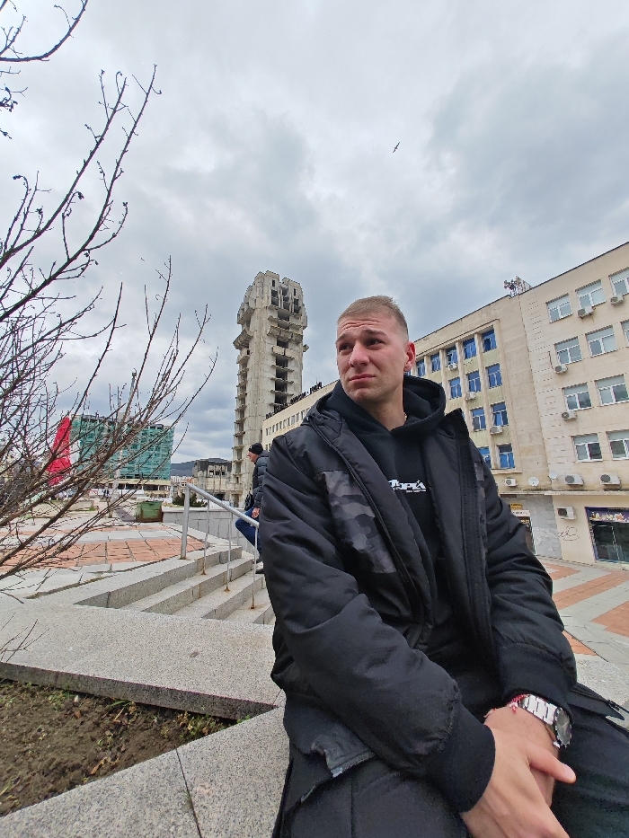 An outdoor, eye-level shot in Shumen, Bulgaria, depicts a young adult male seated on a concrete ledge in the foreground. He possesses fair skin, short light-colored hair, and a pensive expression, with his gaze directed upward and to the right. He wears a black hooded sweatshirt beneath a black winter jacket featuring subtle dark camouflage patterns on the shoulders, and a silver-toned wristwatch on his left wrist. His hands are clasped.
In the mid-ground, to the left, stands a tall, unfinished concrete Brutalist structure, identifiable as the "Clock Tower" or "New Municipality Building" in Shumen. To the right of this tower is a multi-story building with a light-colored facade, numerous windows with blue frames, and external air conditioning units. Further left, another building with a green-tinted glass facade and a large red banner is visible. A set of concrete steps with metal railings descends behind the primary subject, where another male figure is partially observed. Bare tree branches are prominent in the upper left foreground. The sky is overcast with gray clouds, and a small bird is visible in flight in the upper right quadrant. The ground consists of patterned paving and small patches of soil and sparse vegetation.