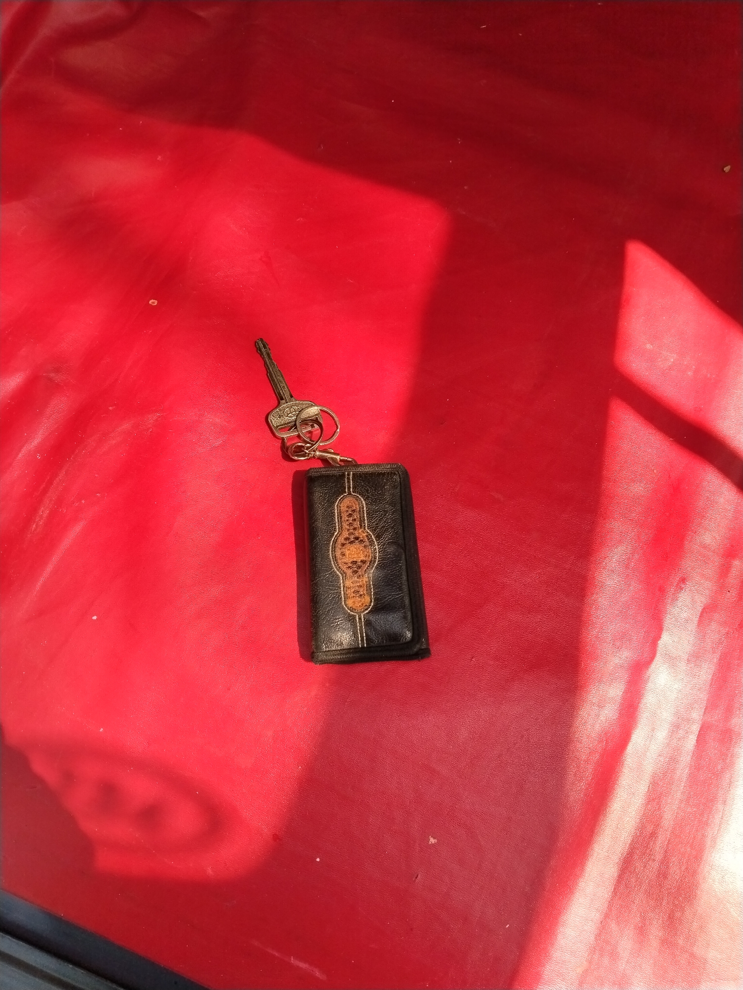 A single car key with a black leather keychain is lying on a red textured surface, possibly vinyl or leather, in what appears to be an outdoor setting in Prigi, Indonesia. The surface exhibits strong sunlight and shadow patterns, indicating a sunny day. The key has a silver-colored metallic head with an engraved pattern and a silver ring attached. The keychain is rectangular, black with gold stitching along the edges, and features an ornate, oval-shaped design in the center with a tan, patterned insert. There are a few small white specks scattered across the red surface, possibly dust or debris. The overall impression is of an everyday object left momentarily unattended on a vibrant, sun-drenched surface.