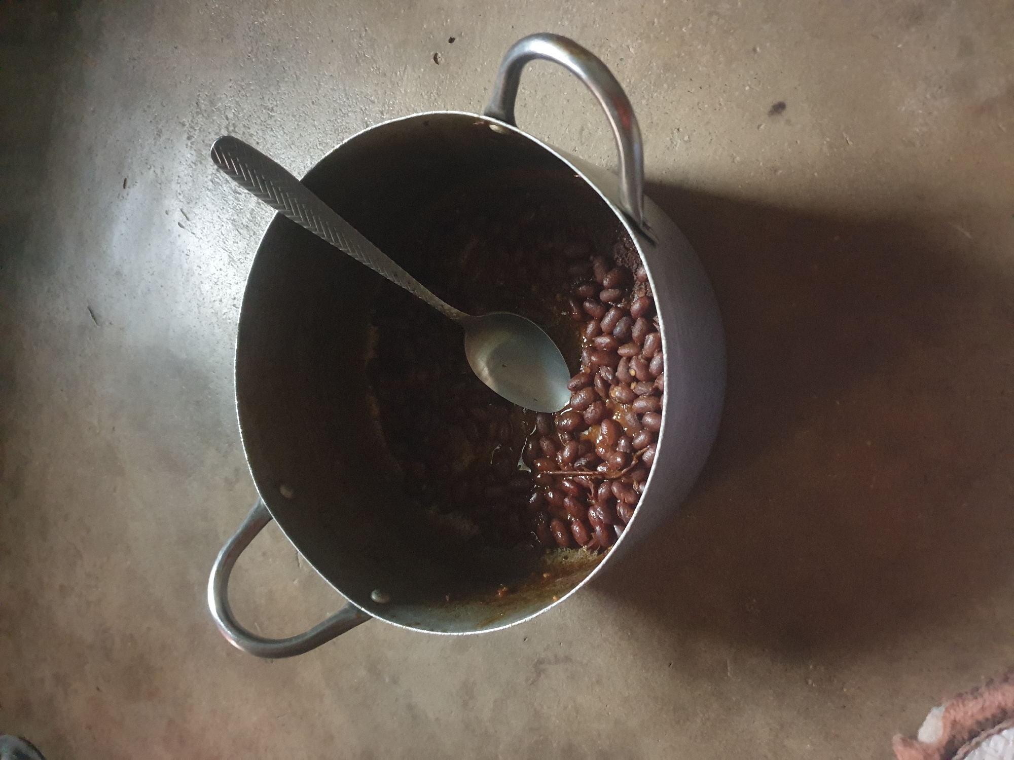 A pot of kidney beans with a spoon rests on a concrete floor. The pot is silver with two handles and contains cooked kidney beans in a dark sauce. The spoon is also silver with a textured handle and is partially submerged in the beans. The concrete floor is a light brown color and appears to be outdoors or in an unfinished space. The lighting suggests it is daytime.
