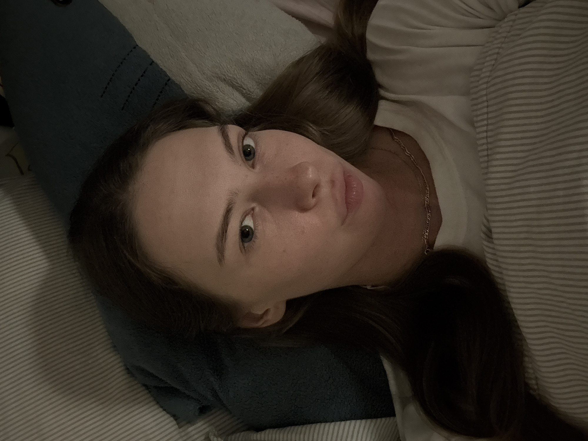 A young woman is lying in bed, looking at the camera. She has light brown hair and green eyes. She is wearing a white t-shirt and a necklace. The background consists of pillows and bedding. The lighting suggests it is nighttime or indoors with dim lighting. The scene is a bedroom. The provided context does not mention or suggest the image is in the city of Perm, Russia.
