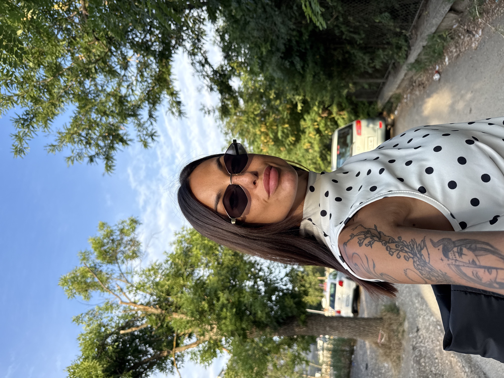 A medium shot captures a woman with long, dark hair and sunglasses, wearing a white polka dot top. She is looking towards the camera with parted lips. Her left arm, visible in the foreground, is adorned with intricate black tattoos, including floral designs and a portrait of a woman with horns. The background depicts an outdoor scene with trees, a dirt path, and two cars parked on the side. The sky is blue with scattered clouds. The image appears to be taken on a sunny day. The location is likely a street or a path in the city of Pleven, Bulgaria, given the typical architecture and vegetation.