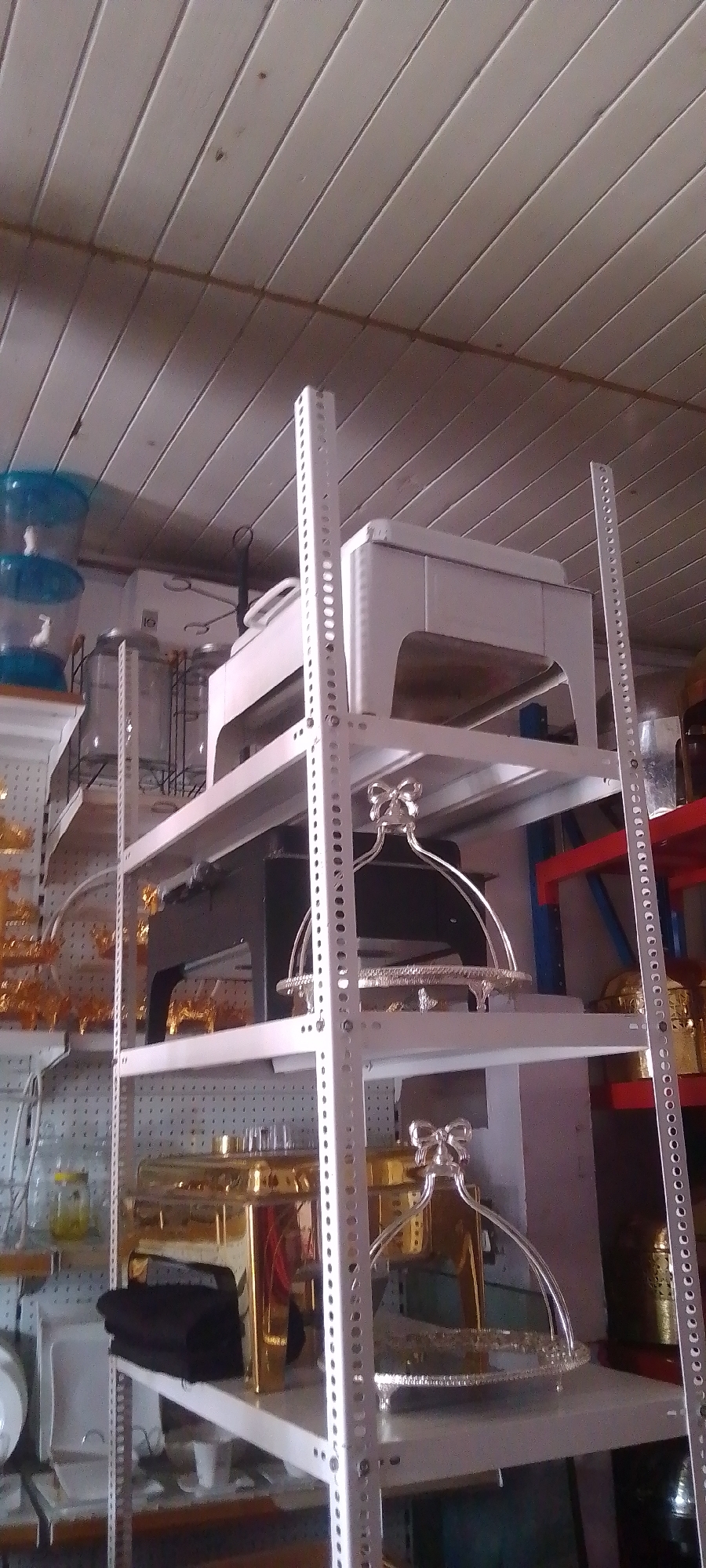 The image shows a metal shelving unit with various objects on it. The shelving unit is made of white metal and has a perforated design. On the top shelf is a white object with a rectangular base and a curved top. Below that, on another shelf, is a black object. On the lower shelves are gold and silver serving dishes. The dishes have decorative handles and ornate designs. The background is cluttered with various other objects, including some blue containers on the left side of the frame. The setting appears to be in a storage area or warehouse. The location is Unknown, Unknown.