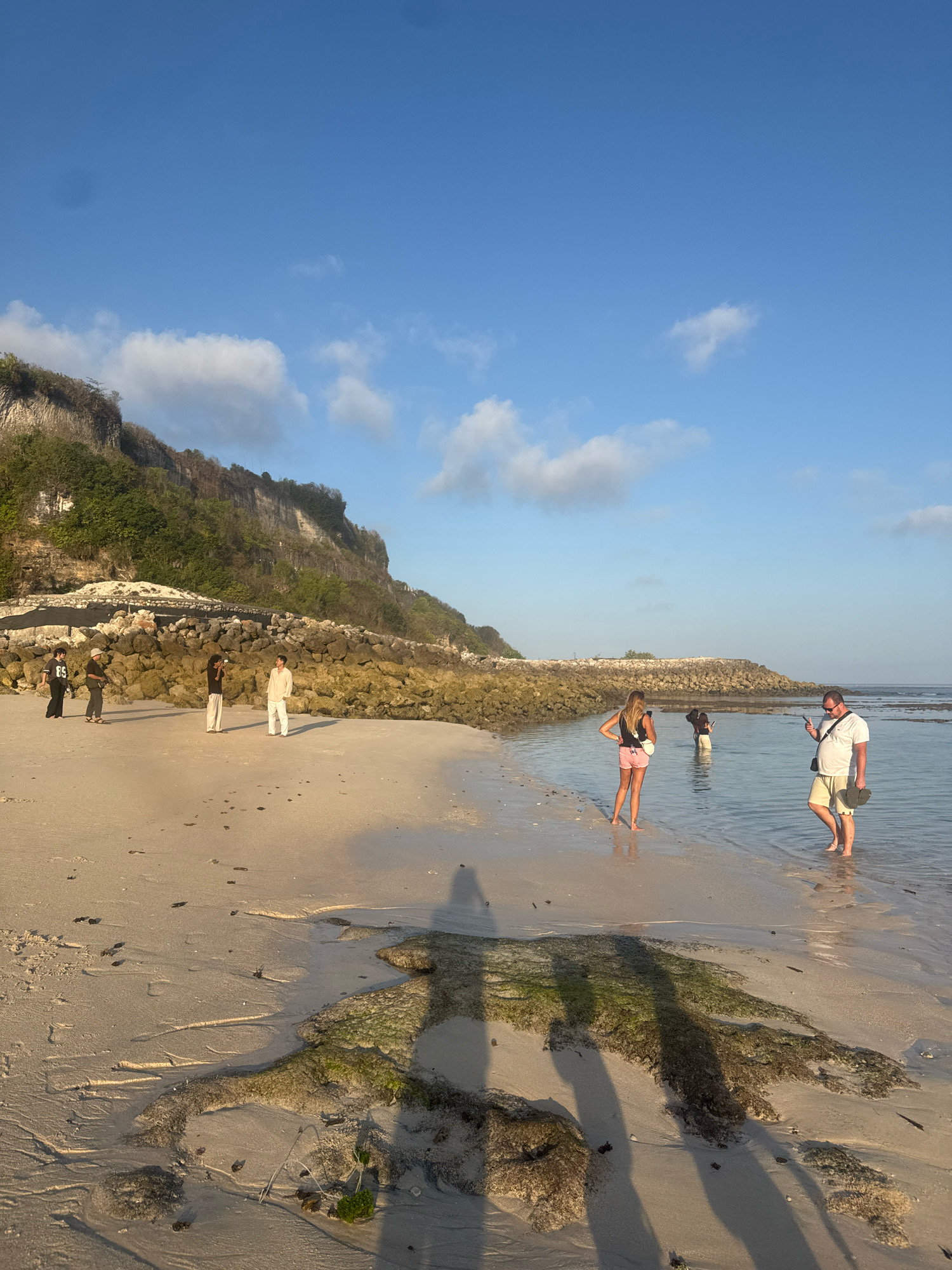 A group of people are gathered on a sandy beach in Kuta Selatan, Indonesia, with a rocky cliff face and the ocean in the background. The sun is low in the sky, casting long shadows. The beach is wet and littered with small pieces of debris. The water is shallow and clear, with some rocky outcrops visible. The sky is blue with a few scattered clouds.
On the left side of the image, four individuals stand on the dry sand, facing the ocean. They are dressed casually, with one person wearing a black shirt with graphics, another in a brown long-sleeved shirt and light-colored pants, and two others in light-colored shirts and pants.
Further down the beach, closer to the water, a group of three individuals are wading in the shallows. Two women, one with blonde hair wearing a black top and pink shorts, and another with dark hair wearing a black top and white pants, are facing away from the camera. A man, wearing a white t-shirt, khaki shorts, and sunglasses, stands to the right, holding a phone and a pair of flip-flops.
The background features a rugged, vegetated cliff face, likely part of the coastline. A wall of large, rough-hewn rocks forms a barrier along the edge of the beach, possibly for erosion control. The ocean stretches out to the horizon, appearing calm and serene. The overall scene depicts a relaxed, seaside gathering at what appears to be a natural coastal area.