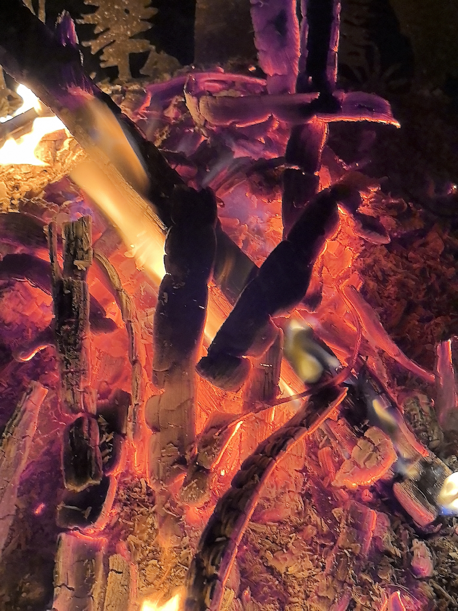 The image depicts a close-up view of a campfire with glowing embers and burning logs. The flames are a vibrant orange and yellow, with purple hues present in the glowing coals. The wood is charred and broken, showing the process of combustion. The background is dark, suggesting it is nighttime. There are no people or specific urban landmarks visible that would indicate a location in Taldykorgan, Kazakhstan. The scene is a natural phenomenon of fire and burning wood.