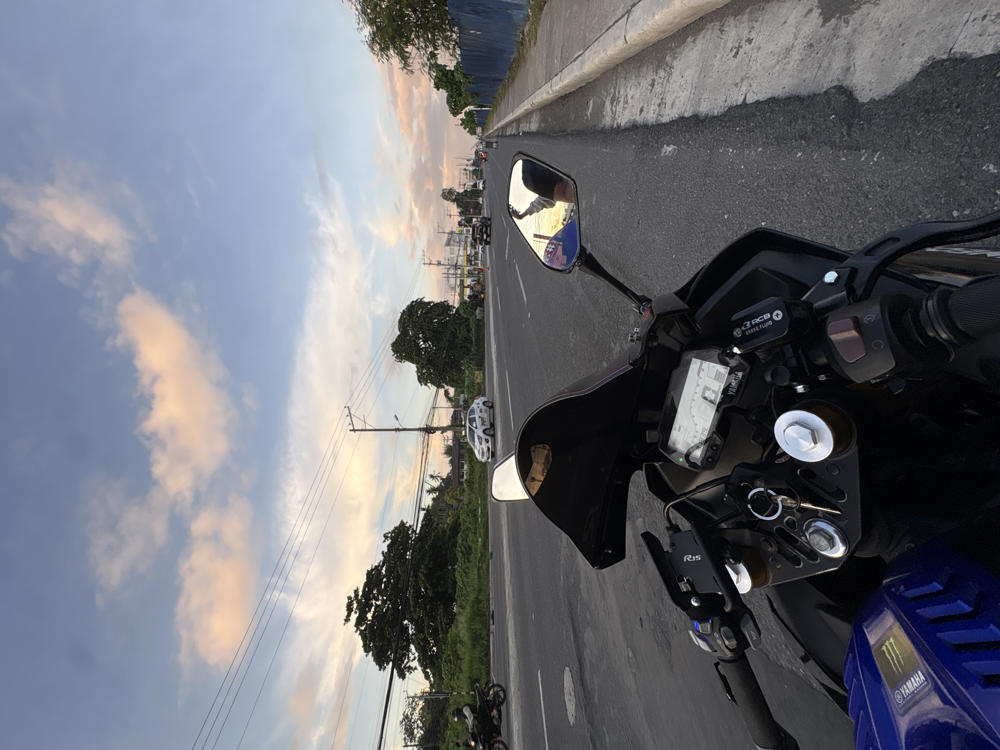 This is a first-person perspective photo of a motorcycle parked on the side of a road in Davao City, Philippines, at dusk. The sky is a gradient of blue to pink and orange, with scattered clouds. The road is asphalt, with a concrete curb and sidewalk on the right. Vegetation, including trees and bushes, lines the left side of the road. Power lines and utility poles are visible in the background. In the foreground, the motorcycle's handlebars, dashboard, and part of the fuel tank are visible. The dashboard displays digital readouts. A set of keys is in the ignition. A side mirror reflects the sky and the back of the rider's head and shoulder. Traffic is visible on the road, including cars and other motorcycles. The overall scene suggests a moment of pause during a motorcycle ride during sunset in an urban or suburban environment.