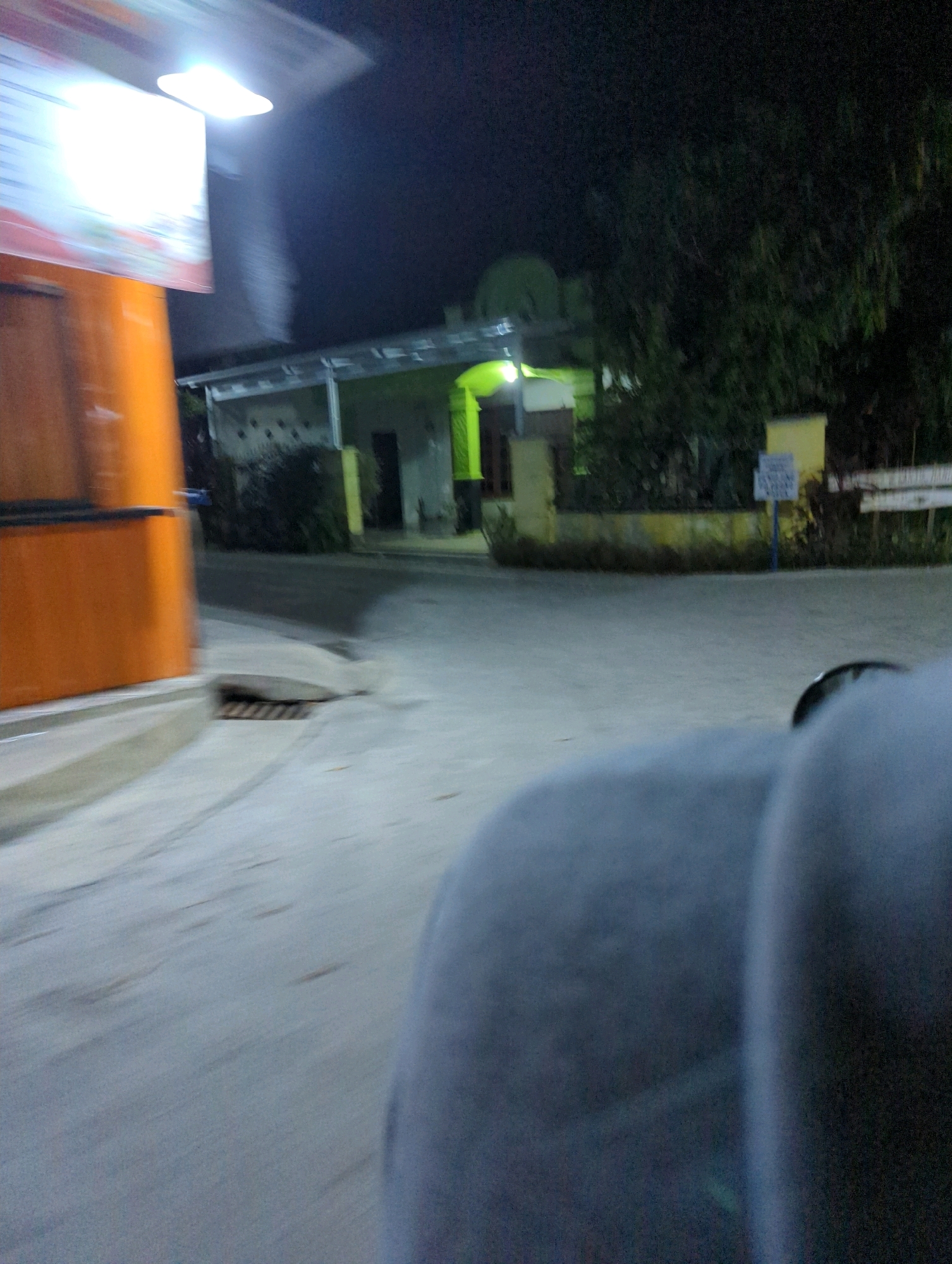 This is a nighttime, blurry, exterior photograph taken from a moving vehicle in Senggrong, Indonesia. The scene shows an orange structure on the left, possibly a food stall or kiosk, with a bright illuminated sign above it. To the right of the orange structure, there is a grey concrete pathway and a drain grate. Further in the background, there is a building with a green roof and yellow accents, surrounded by trees. The ground appears to be asphalt or concrete, and there are some signs visible near the building. The foreground is dominated by a blurry grey object, which is likely part of the vehicle the photograph is being taken from, such as a seat or person's clothing.
