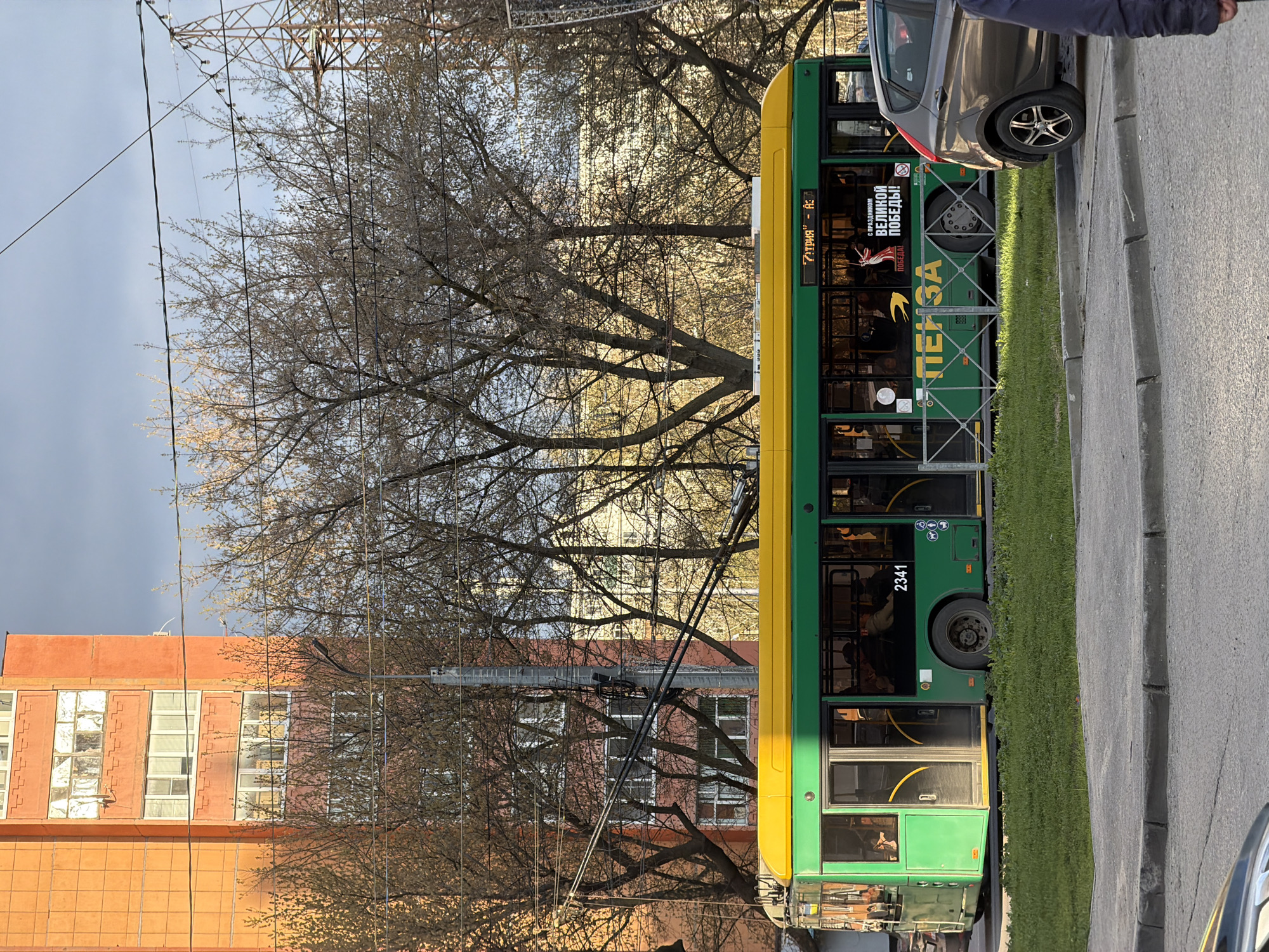 This image depicts a daytime street scene in Penza, Russia, centered around a green and yellow trolleybus. The trolleybus, bearing the number "2341" on its side, is stopped or parked along the road. Its overhead poles are connected to visible power lines that crisscross the sky. Several passengers are discernible inside the vehicle through its windows.
To the right of the trolleybus, a partial view of a gray car is visible, with a person's arm or jacket also in the frame. On the left, a large, multi-story building with a reddish-orange facade and numerous windows stands behind the trolleybus. The background is filled with many bare deciduous trees, indicating late winter or early spring. Overhead, beyond the trees and buildings, a partly cloudy sky is visible.
Notable text on the side of the trolleybus includes "ТЕЙ ЗА" (possibly part of a larger word or advertisement), and a prominent banner proclaiming "С ПРАЗДНИКОМ ВЕЛИКОЙ ПОБЕДЫ!" (Happy Great Victory Day!), a clear seasonal cue pointing to late April or early May. Another fragment "ГАТРИЯ" - А:" is also present. The scene captures a moment of ordinary urban transit, reflecting a calm, everyday atmosphere.