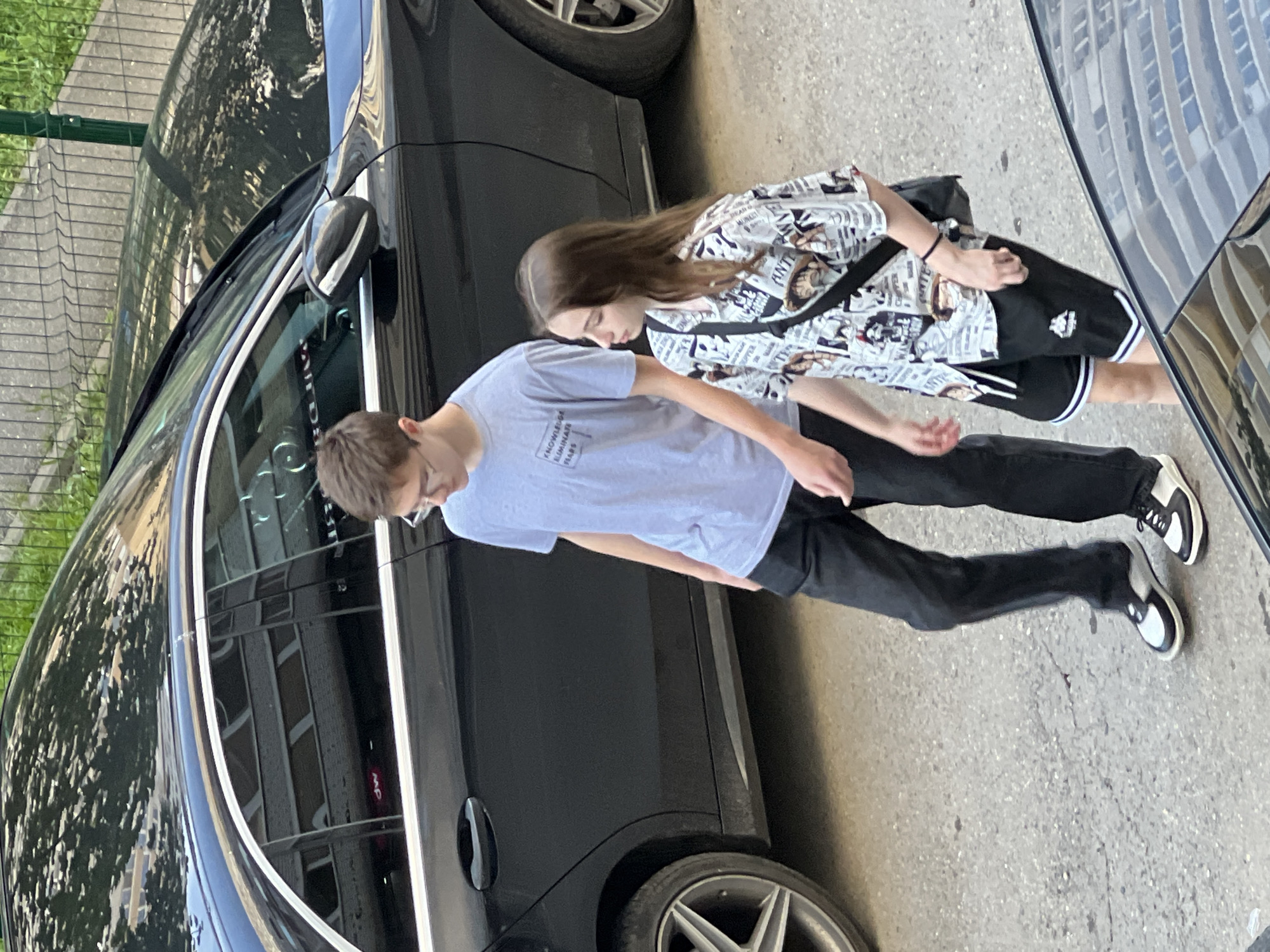 Two young individuals are captured walking or standing next to a parked black car on what appears to be an asphalt surface, possibly a street or parking lot, in Krasnodar, Russia. The image is taken during the daytime, indicated by bright lighting and visible shadows, suggesting a sunny day.
The subject on the left is a male with short brown hair, wearing glasses, a light grey t-shirt, and dark pants. His t-shirt clearly displays the text: "KNOWLEDGE ELIMINATES FEARS". He is also wearing black and white sneakers. He is looking downwards.
The subject on the right is a female with long brown hair. She is wearing a patterned short-sleeved shirt that resembles newspaper print, black shorts with white stripes along the hem, and black and white sneakers similar to the male's. She is carrying a black shoulder bag. Parts of her shirt show text, with "ANTON" visible on one section, and other words like "WORLD" and "DAILY" hinted at amidst the newspaper clippings. Like the male, she is also looking downwards.
A sleek black car, likely a sedan, is parked parallel to them, occupying a significant portion of the left side of the frame. Reflections in the car's dark, glossy windows show what appears to be a multi-story building with numerous windows, hinting at an urban setting. A green wire fence is visible in the upper left background, behind the car, suggesting a boundary or a patch of green space. The overall scene depicts a casual outdoor moment.
