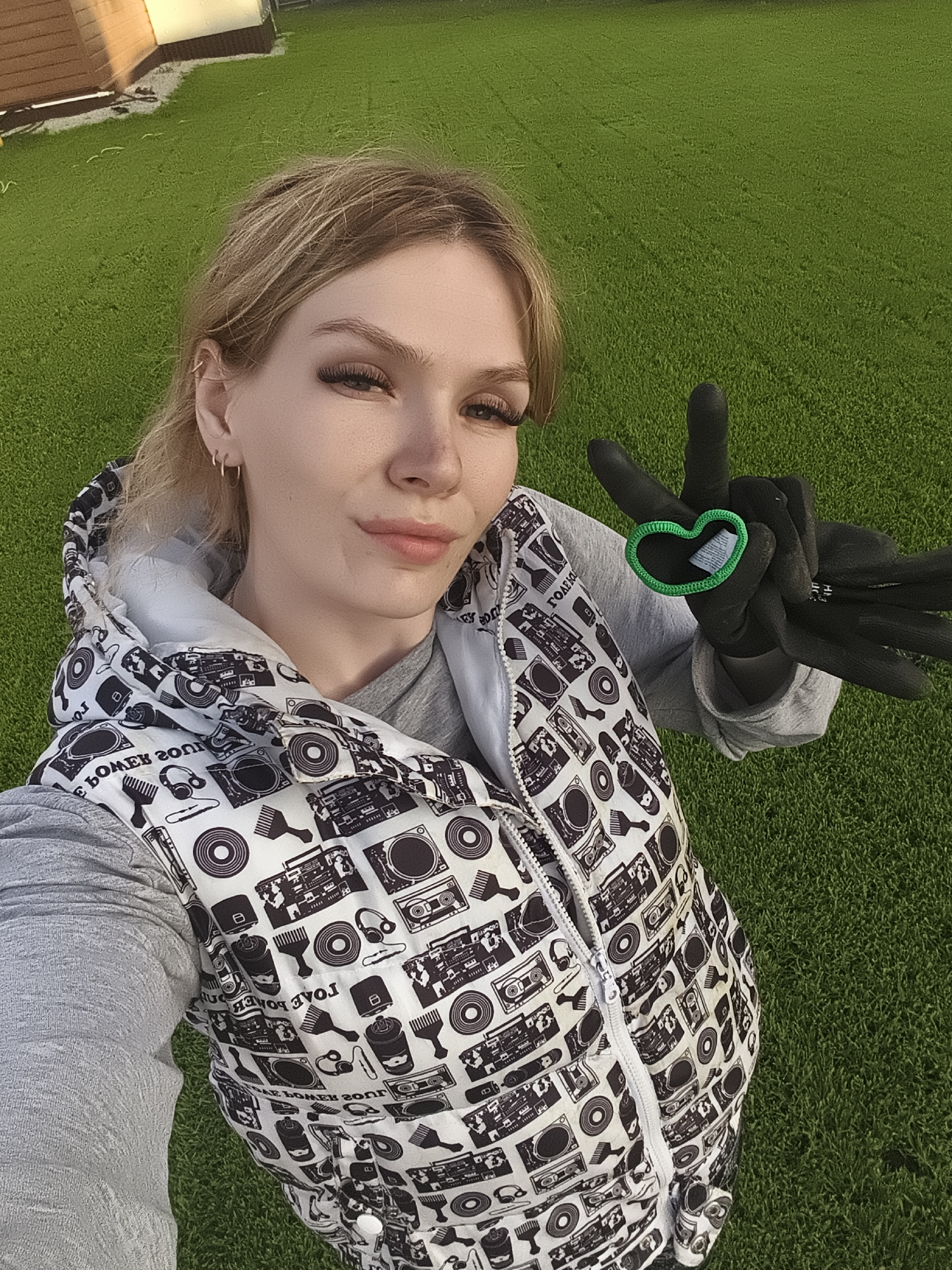 A young woman is captured in a selfie taken outdoors in Русский Вожой, Russia. She has blonde hair, pulled back, and is looking directly at the camera with a slight, confident smile. Her eyes feature prominent false eyelashes.
She is wearing a gray long-sleeved shirt beneath a white and gray hooded vest. The vest is patterned with black and white illustrations of various music-related items, including boomboxes, records, headphones, combs, and spray cans. Mirrored text such as "I LOVE POWER" and "BOOM BOX" is visible within the pattern on her vest due to the selfie orientation.
Her left hand, adorned with a black glove, is raised, forming a peace sign. Between her index and middle fingers, she holds a bright green heart-shaped object, possibly a hair tie or small decorative item.
The background shows a uniformly green, well-maintained grassy area, which appears to be artificial turf. To the upper left, a wooden structure, possibly a shed or part of a building, is visible, with its upper section painted a lighter color. The area is brightly lit by sunlight, suggesting a clear day, likely late afternoon or early evening given the lighting angle on the background structure. The overall scene conveys a casual and pleasant outdoor moment.