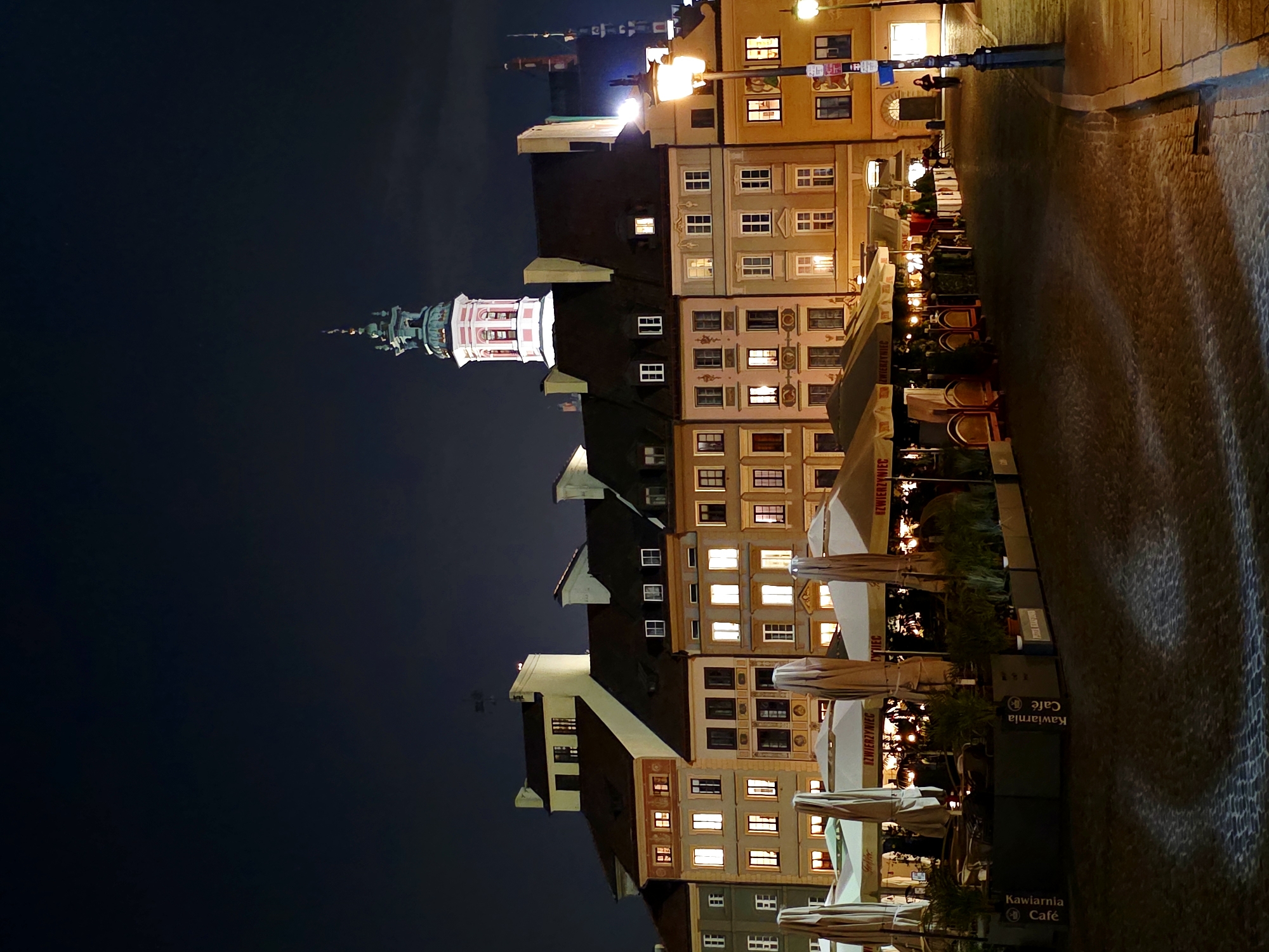 The image depicts a nighttime view of the Old Town Market Square in Warsaw, Poland. In the foreground, several outdoor cafe seating areas are visible, covered by large umbrellas and surrounded by planters containing greenery. The cobblestone ground is illuminated by streetlights and the warm glow from the windows of the surrounding historic buildings. The buildings themselves feature colorful facades and ornate architectural details, typical of Warsaw's reconstructed Old Town. Prominently featured is a church tower with a distinctive green spire, lit up against the dark night sky. One or two figures can be seen walking along the street. The scene conveys a lively yet peaceful atmosphere characteristic of a European city square at night.
