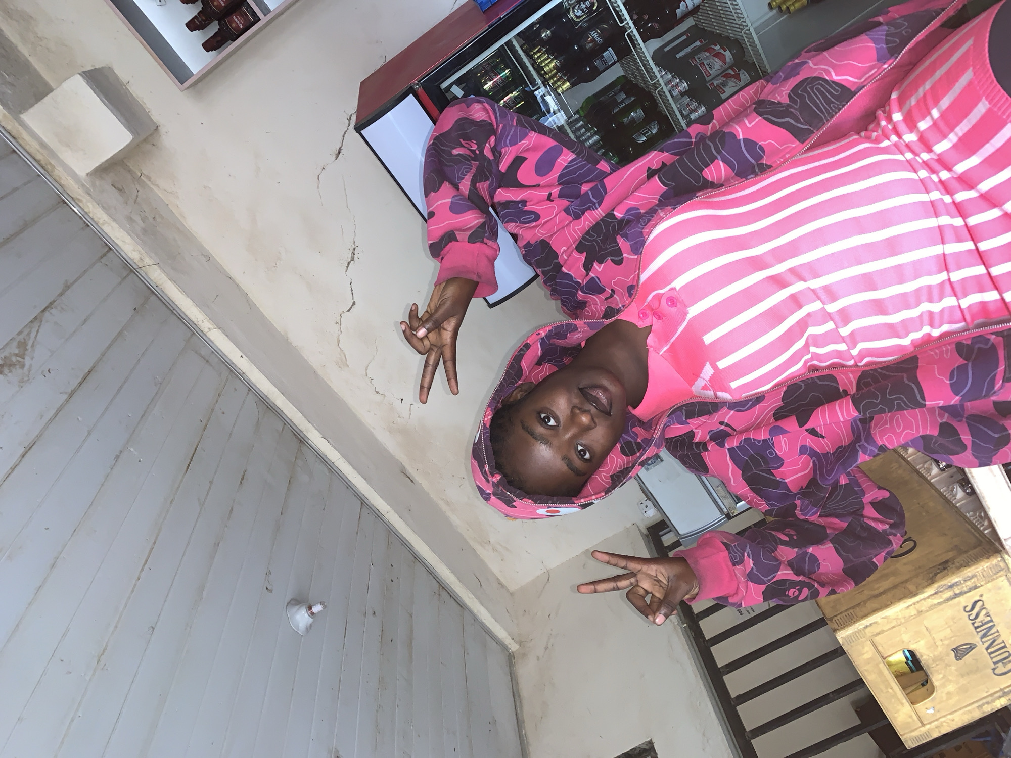 A young Black woman is pictured in a close-up, medium-angle shot, posing with a peace sign gesture. She is wearing a vibrant pink hooded jacket with a camouflage pattern in shades of pink and purple, layered over a pink and white striped dress. The background shows a cluttered interior space, possibly a small shop or storage area. To the left, a wooden crate with the "Guinness" logo is visible, and to the right, a red beverage cooler is stocked with bottles and cans. The ceiling, made of white wooden planks, is visible at the top of the frame, with a light fixture attached. The overall impression is a candid snapshot taken in an informal setting in New Stadium, Nigeria.