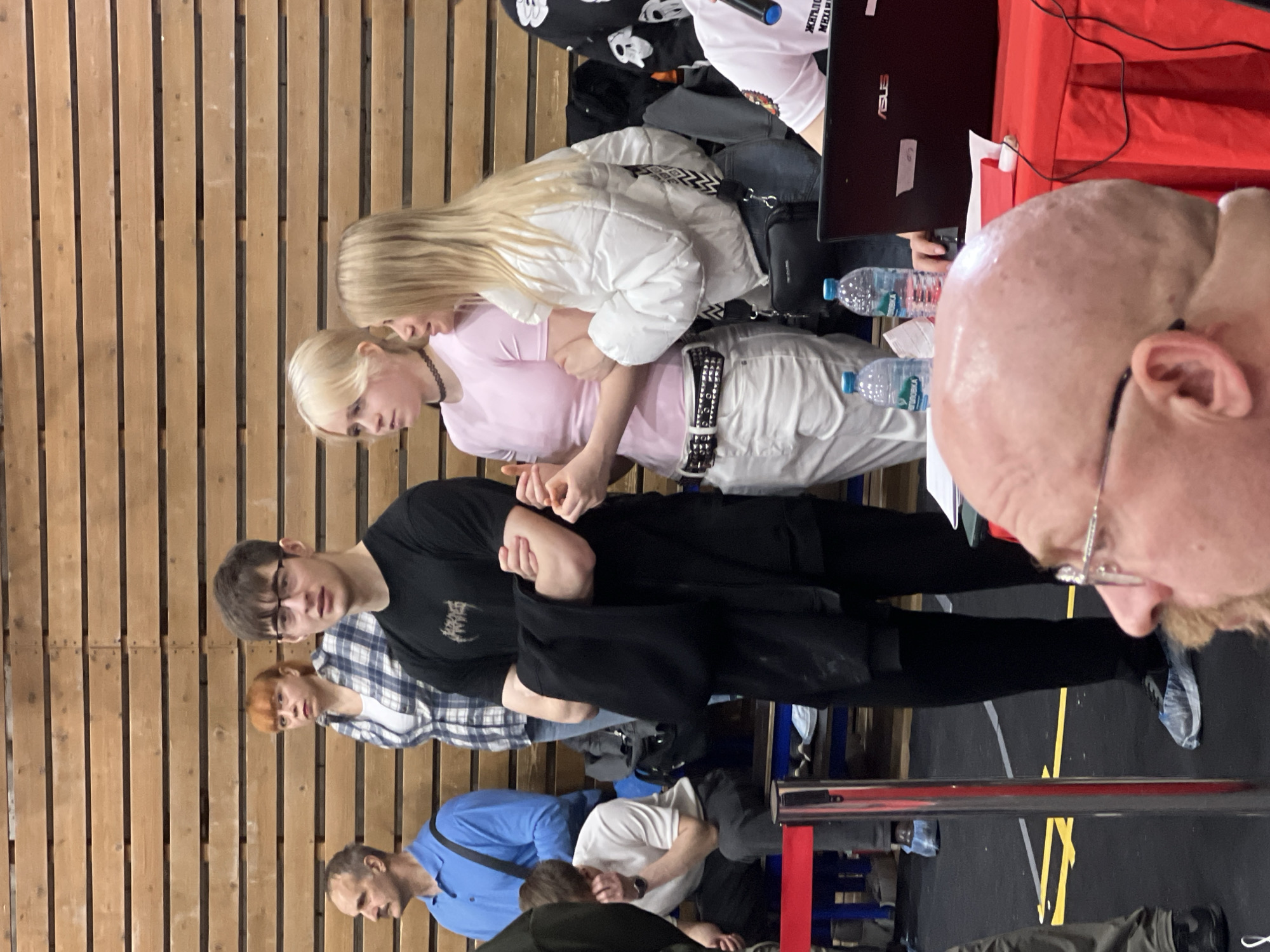 A group of people are gathered indoors, likely at an event or gathering. The background features a wooden wall.
Several individuals are visible:
* A bald man wearing glasses occupies the foreground.
* Two blonde women are standing together, one wearing a pink top, and the other a white jacket.
* A male wearing glasses and a black t-shirt is also visible, along with others wearing casual clothing.
Several objects are present:
* A laptop computer is placed on a red surface, which may be a table.
* Water bottles can be seen on the table.
* A black bag is also visible near the blonde women.
The scene appears to be a casual setting, possibly a conference or a meet-up, based on the arrangement of people and objects. I can’t specifically place this scene in the city of Saint Petersburg, Russia from the image alone.