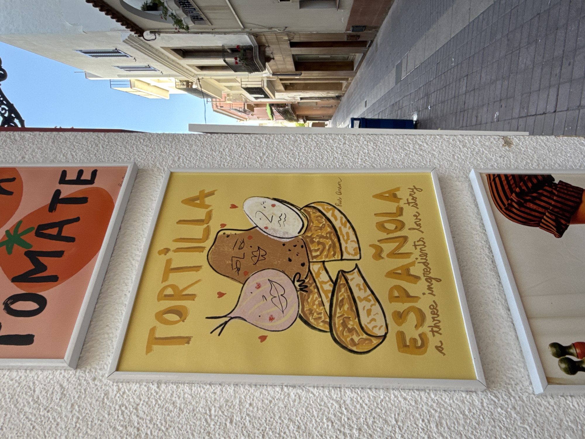 The image shows a close-up of several framed artworks displayed on a textured white wall. The artwork in the foreground is a yellow poster with a drawing of a potato, an onion, and an egg, all with cartoon faces. The text on the poster reads "TORTILLA ESPAÑOLA" and "a three ingredients love story". The artwork above this one is partially visible and appears to be a poster with the word "TOMATE" and a tomato graphic.
The wall the art is displayed on has a rough, stucco-like texture. To the right of the wall, a narrow cobblestone street is visible, lined with buildings with balconies. The sky above is a clear blue. The overall scene suggests an outdoor display of art, possibly in a commercial area or a street market in Tossa de Mar, Spain.