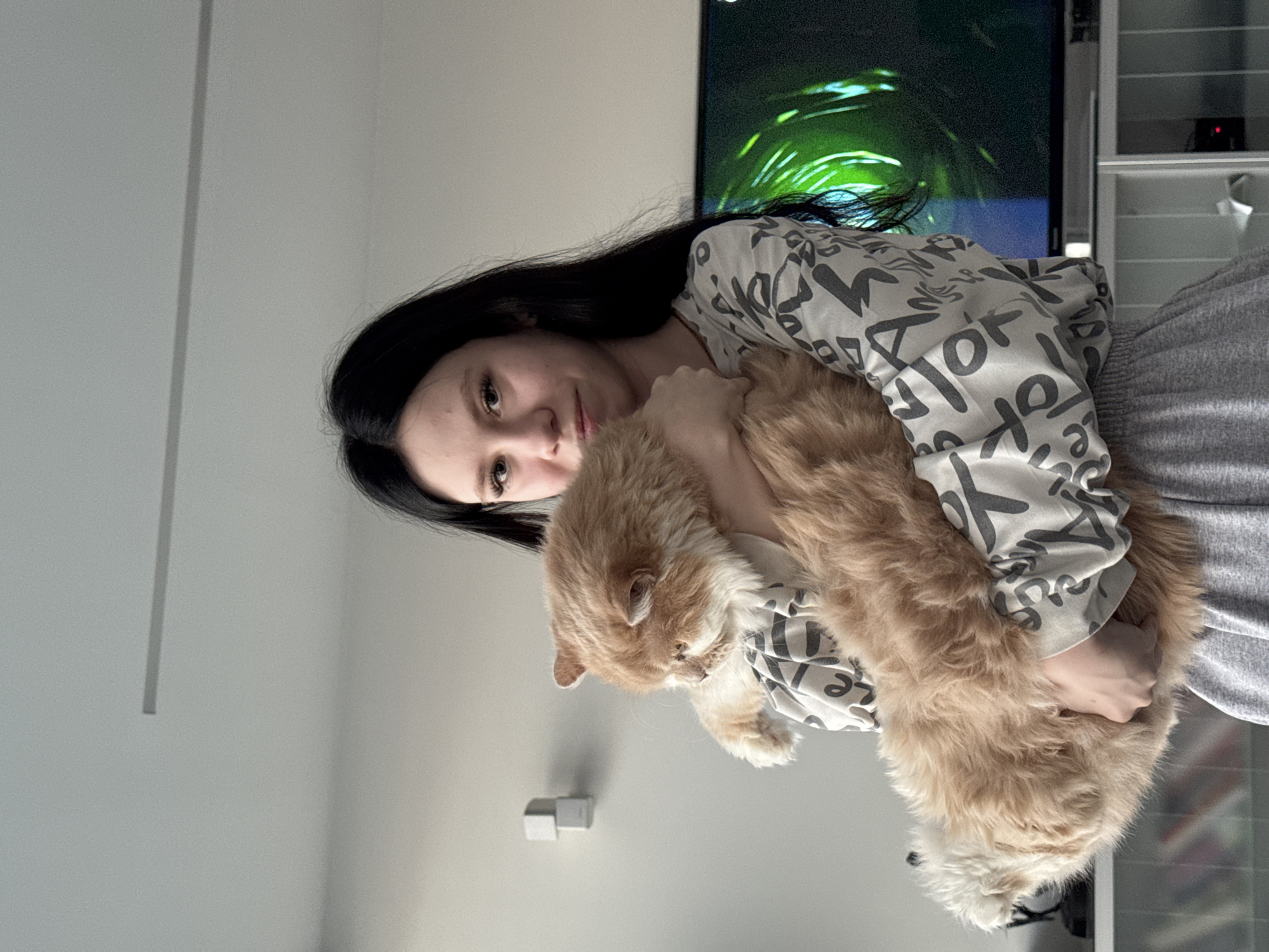 A woman with long, dark hair is holding an orange cat. She is wearing a white shirt with gray lettering and gray sweatpants. The background includes a white wall, a television with a green screen, and a white shelf. The location is unknown.