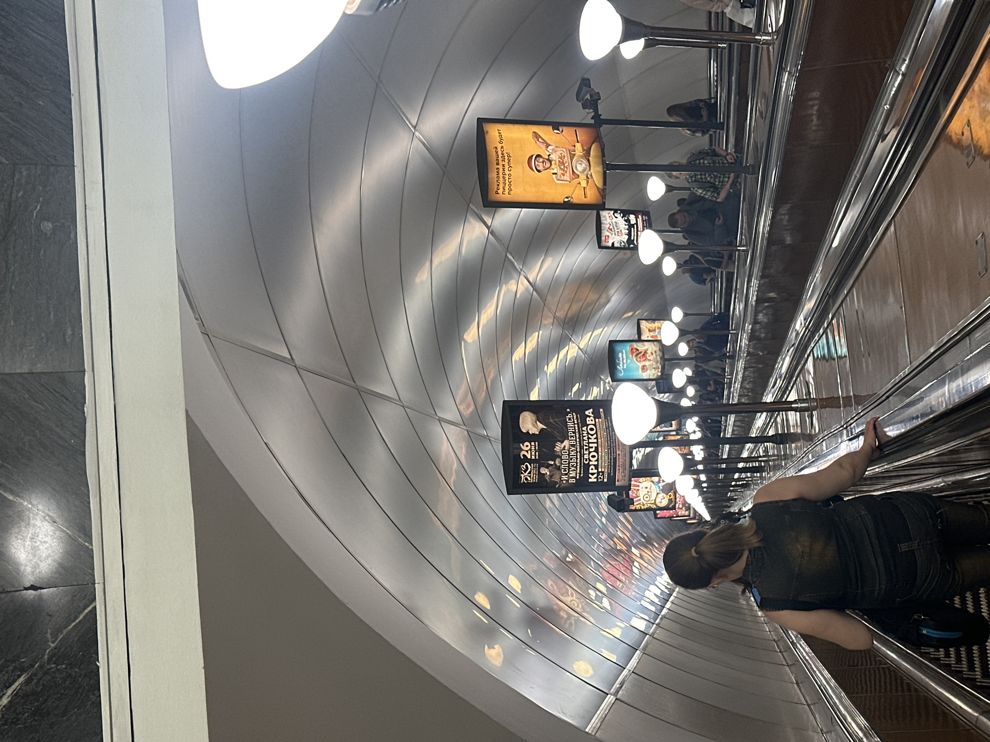The image shows a woman from behind, riding an escalator in a metro station. She is wearing a dark sleeveless vest over a dark shirt and dark pants, and has her hair in a ponytail. She is holding onto the handrail of the escalator with her right hand. The escalator is lined with lights and advertisements on the curved ceiling. One advertisement, prominent in the center-left, features a poster for a performance titled "И СЛОВО В МУЗЫКУ ВЕРИШЬ" by Светлана Крючкова. Another advertisement on the right side of the escalator depicts a man on a scooter with a pizza box, with the text "Реклама вашей пиццерии здесь будет просто супер!". Other people are visible on escalators going in the opposite direction and on the platform. The environment appears to be a modern and well-lit metro station.