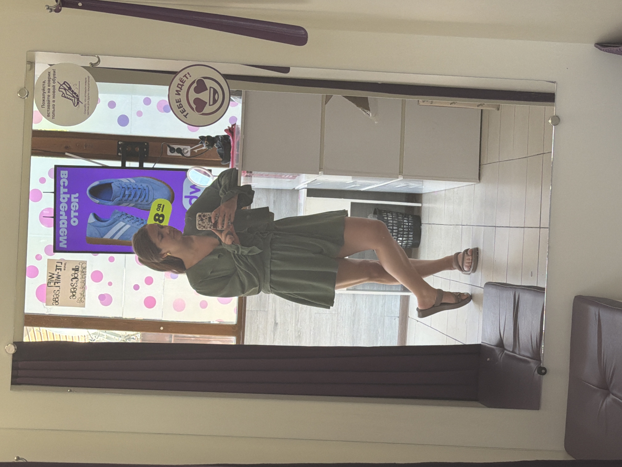 In Sochi, Russia, a young woman is taking a mirror selfie in what appears to be a fitting room, possibly at a shoe store. She is wearing a green, short-sleeved V-neck dress with a tie waist, and brown flat sandals. The woman has long brown hair pulled back into a ponytail and is wearing small gold hoop earrings. She holds a smartphone in her right hand, capturing her reflection. The fitting room features a full-length mirror that takes up a significant portion of the image. Behind her, a purple curtain hangs on the left, and on the right, a display of shoes is visible on a purple background, featuring blue sneakers. A sign with a graphic of shoes and text in Russian reads, "Please, stand on the rug only when trying on new shoes!" Another circular sticker with a smiley face and hearts in its eyes, accompanied by the Russian text "It suits you!", is also present on the mirror. The floor is tiled, and there are white storage units visible. A dark trash can is positioned behind the woman. The overall lighting is bright, suggesting an indoor commercial setting.