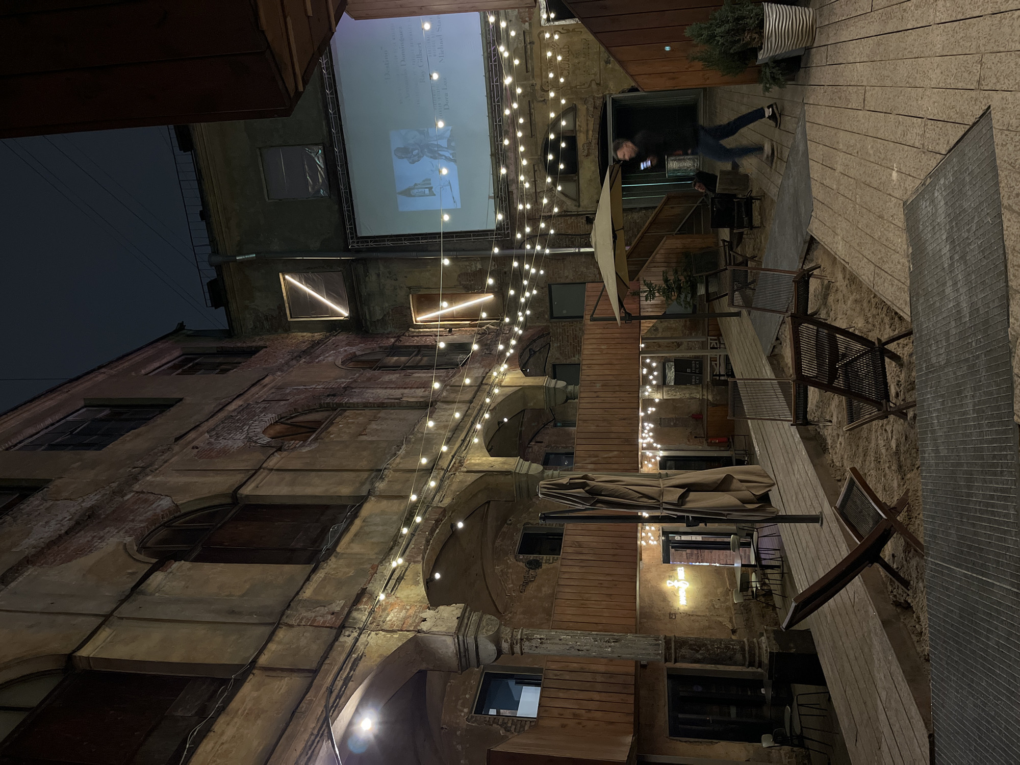 The image depicts an outdoor, illuminated courtyard at night within the industrial-chic repurposed space of Sevkabel Port, Saint Petersburg, Russia. Aged, multi-story brick and stucco buildings, exhibiting signs of historical wear and decay with crumbling plaster and exposed brick, enclose the area. Some windows are boarded or covered, while others feature arched frames.
The courtyard itself features a multi-level layout with elevated wooden walkways and platforms, interspersed with sections of sand. Numerous string lights are strung across the courtyard, illuminating the scene. Various outdoor furniture is present, including lounge chairs placed in the sand, tables with chairs, and a large folded beige umbrella. Potted evergreen plants are positioned throughout the space.
A large projection screen is mounted on one of the buildings, displaying a film or presentation that includes an image of a stylized figure and legible text, notably "Destino" and several names such as "Alexandre Dominguez" and "Michael Starr," suggesting movie credits. A prominent diagonal neon light fixture is visible on a building wall near the screen, and another neon sign reading "Happy B" (partially obscured) is visible above an entrance.
In the midground, a lone male individual, dressed in dark clothing (dark jacket, blue jeans, light-soled sneakers), is captured in motion, walking from right to left across a wooden path. He is holding and looking at a smartphone. The overall ambiance is that of an active, urban outdoor event or recreational space.