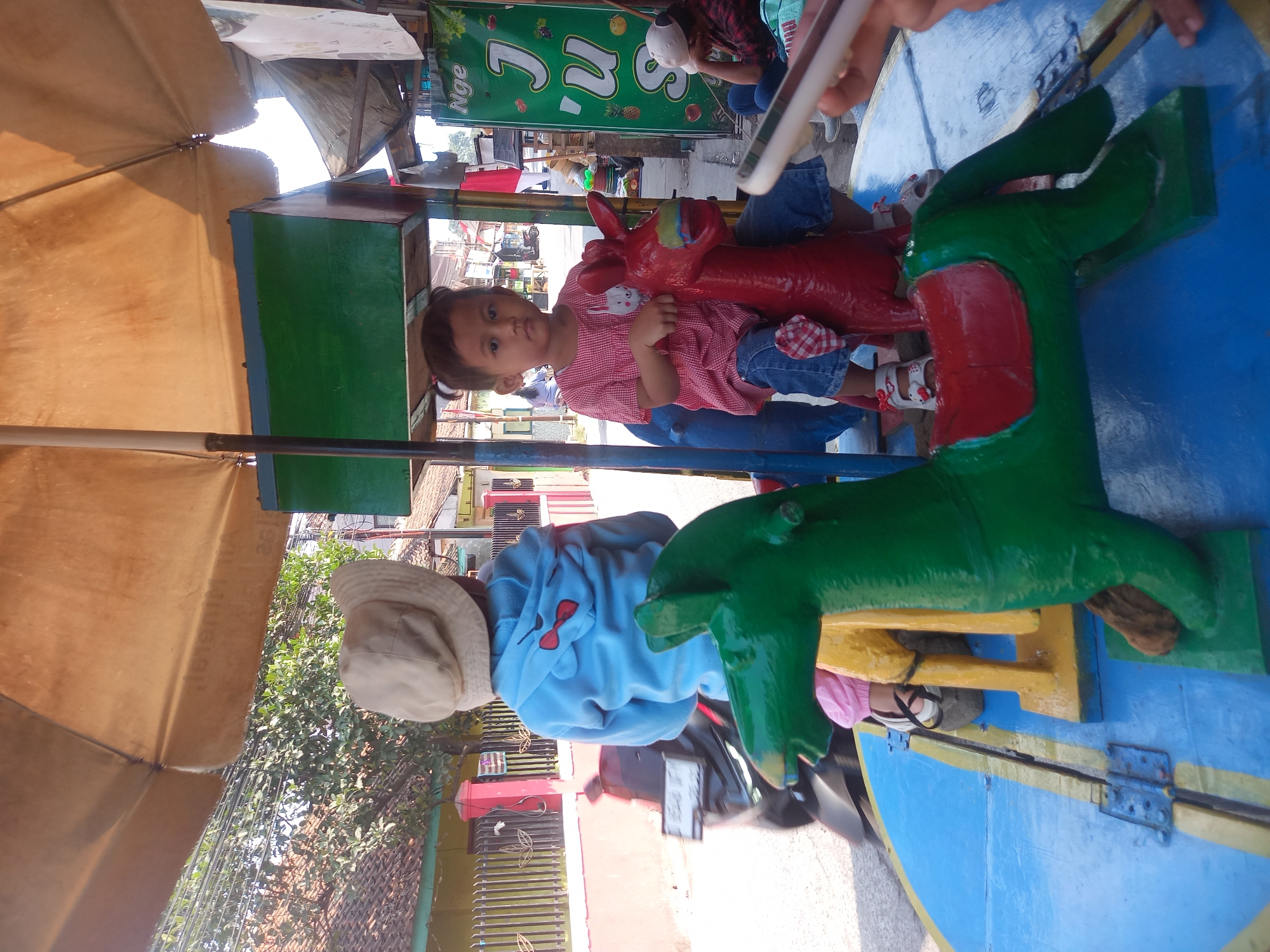 A medium-shot captures two young children seated on a carousel in Linggar, Indonesia. The carousel, featuring brightly painted horses, is positioned outdoors under a large, tan umbrella. The child on the right, a girl with dark hair tied in pigtails, wears a red and white checkered shirt and blue denim shorts. She is mounted on a red horse. The child on the left, partially obscured by the carousel's structure, wears a light blue hooded sweatshirt and a tan bucket hat, and appears to be sitting on a green horse. The carousel itself is painted in bold colors, with a blue base and yellow accents. In the background, a street scene unfolds with houses, a motorcycle, and a sign for "Jus" (juice) suggesting a commercial area. The lighting indicates it is daytime, with sunlight casting shadows. The overall impression is a lively, outdoor recreational setting for children.