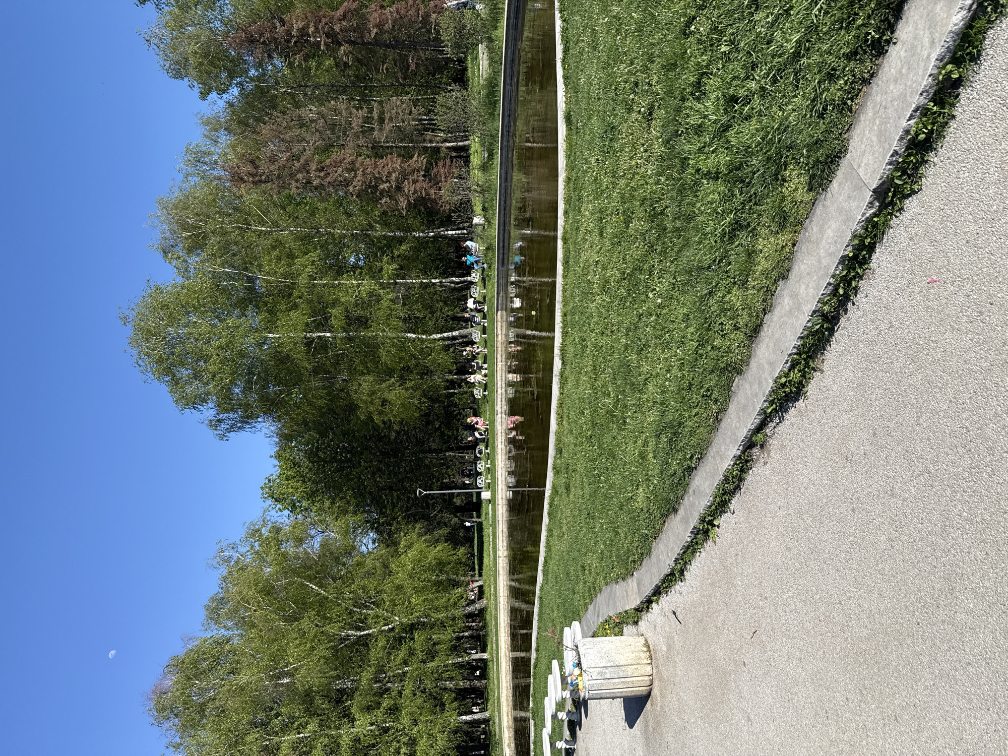 The image shows a park scene with a long, narrow water feature flanked by birch trees and green lawns. People are sitting in white chairs along one side of the water, their reflections visible in the water. A concrete path runs parallel to the lawn. The sky is blue and clear. The moon is visible in the sky on the lower left. It is believed to be located at the National Palace of Culture, Sofia, Bulgaria.