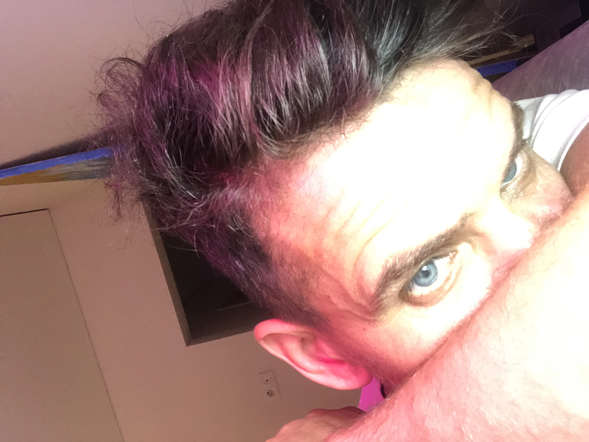 The image is a close-up selfie of a person's face, taken from a low angle. The person has blue eyes and dark hair with streaks of purple. Their head is tilted and their right arm is positioned in front of their face, partially obscuring it. The background is indistinct, with a pinkish wall and what appears to be an electrical outlet on the left. There is no discernible location in Saint Petersburg, Russia.