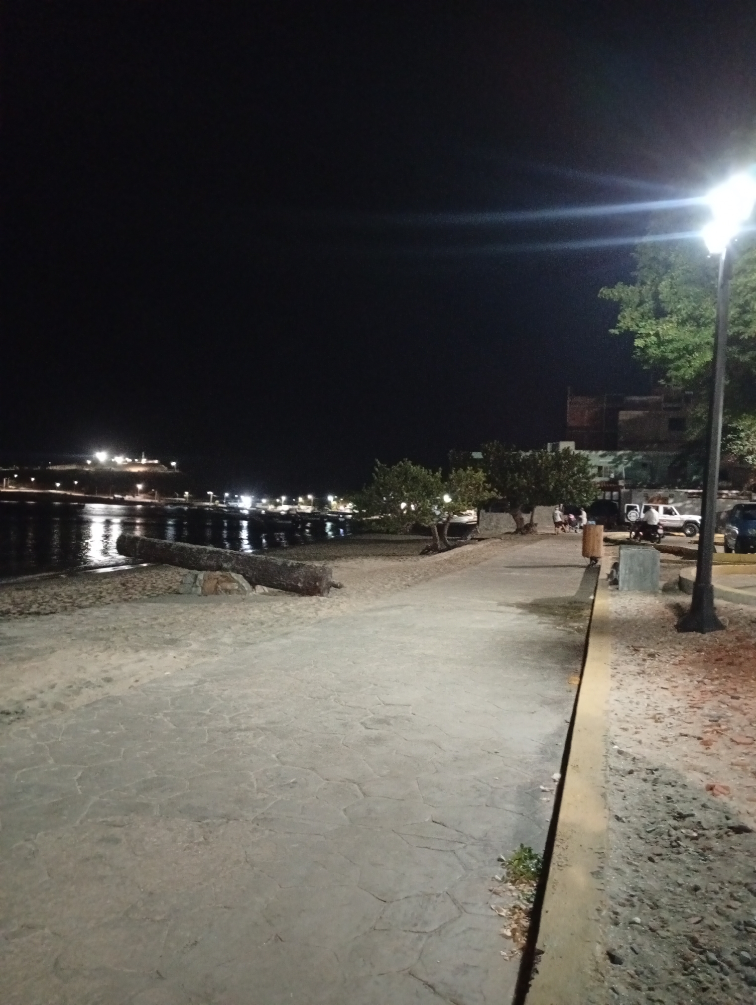 The image depicts a night scene on a waterfront promenade in Juan Griego, Venezuela. The foreground features a textured concrete walkway, bordered by a low, light-colored curb separating it from a sandy area. To the left, there is a beach area with a large, dark object resembling a cannon, positioned near the water's edge.
Across the water, lights from buildings and street lamps are visible on the opposite shore. In the middle ground, trees line the walkway, and further back, a few people are gathered near parked vehicles. A bright street lamp is visible on the right side of the frame, casting a glare. A building is visible behind the trees.