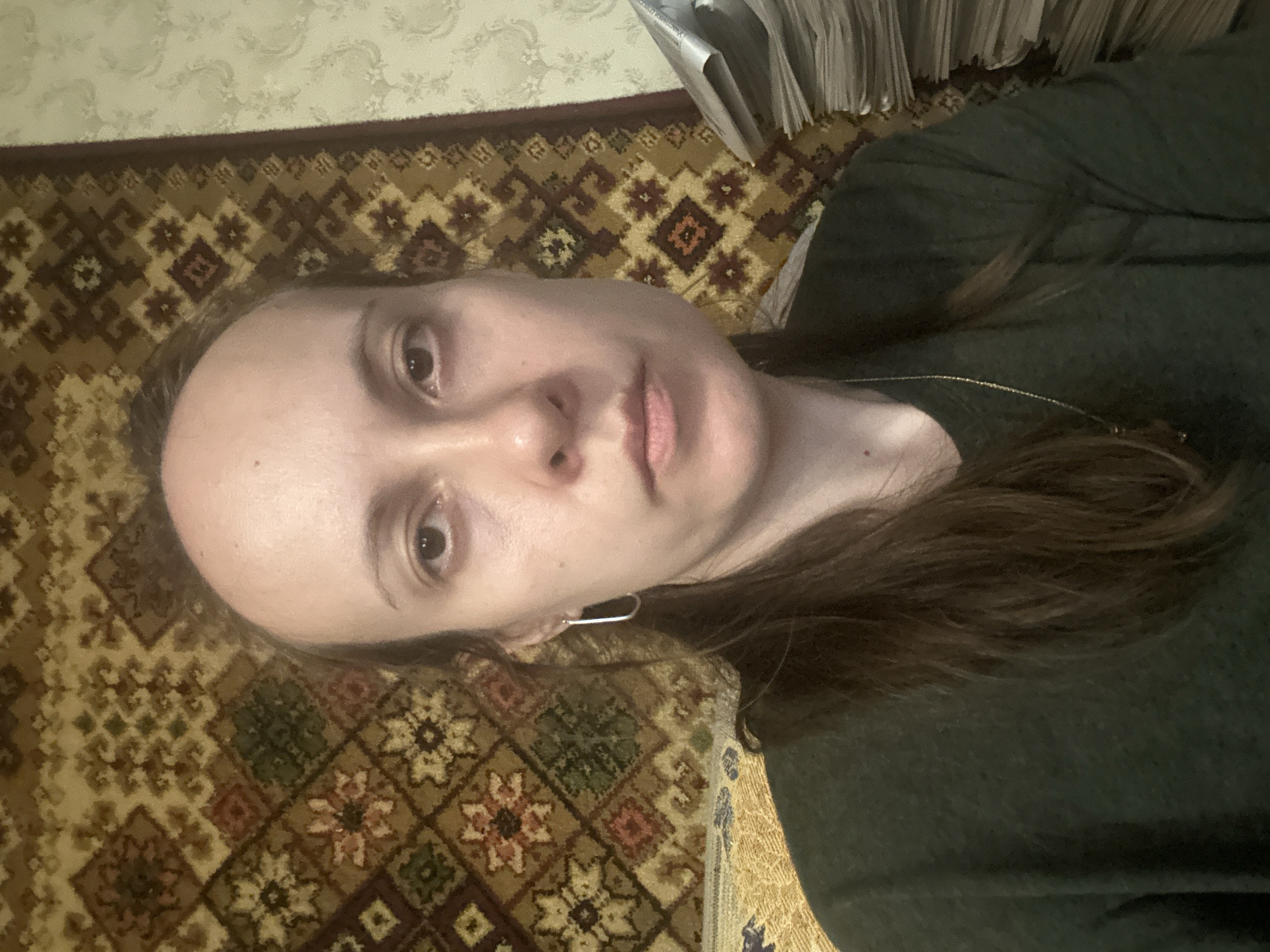 In this head-and-shoulders selfie, a woman with long, dark brown hair tied back, a prominent jawline, and a large lump on the right side of her face is pictured. She is wearing a dark green crewneck shirt and a thin gold necklace. A large, ornate tapestry with geometric patterns in shades of brown, beige, red, and green hangs on the wall behind her. To the right of the woman, a stack of newspapers or magazines is visible. The lighting appears to be artificial and the overall setting is indoors.