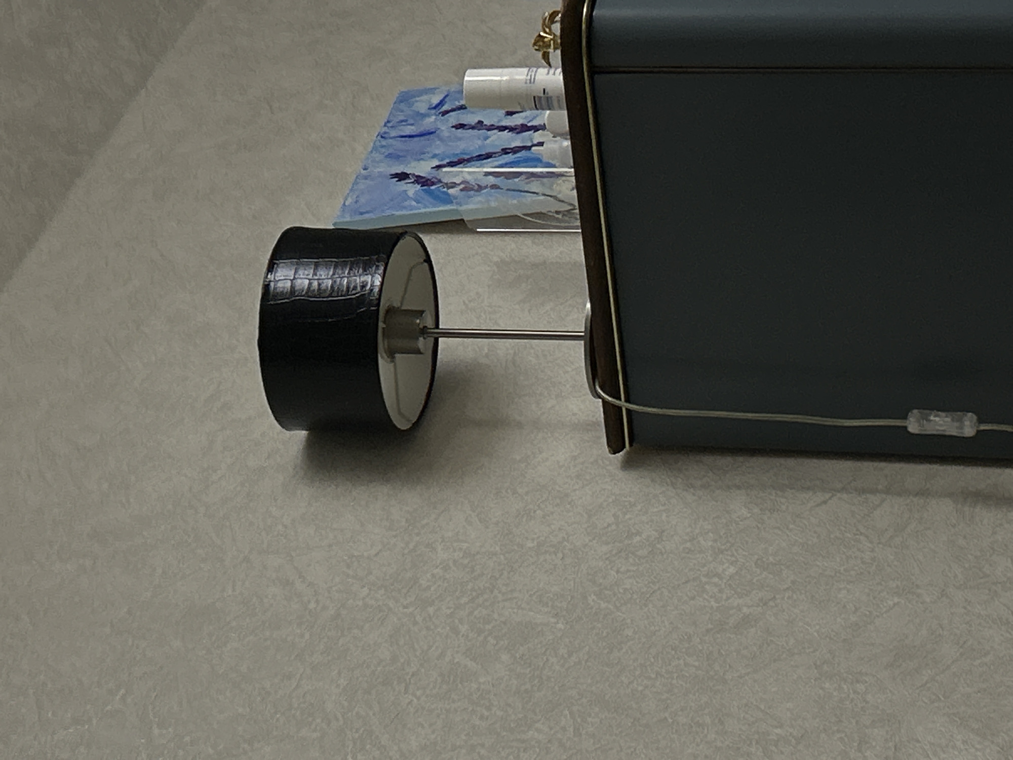 A grey bedside table lamp sits on a grey surface. The lampshade is blue and a black wheel with a silver axle sticks out from the side. On top of the lamp are a small painting and bottles. The location is unknown.