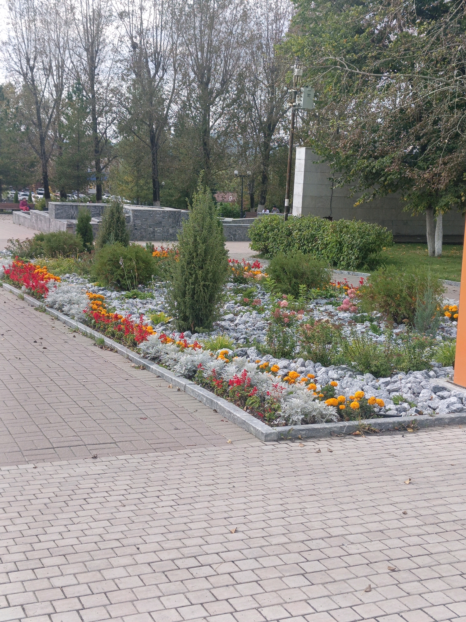 The image captures a paved walkway adjacent to a flowerbed filled with a variety of colorful flowers and greenery. The flowerbed is bordered by grey stones and a concrete edge. In the background, there are trees with sparse foliage, suggesting an autumn or early spring season. Beyond the trees, concrete structures and a large building with a white panel are visible. The walkway itself is made of light-colored paving stones arranged in a pattern. The scene appears to be in a public area, possibly a park or a square, in the city of Satka, Russia.