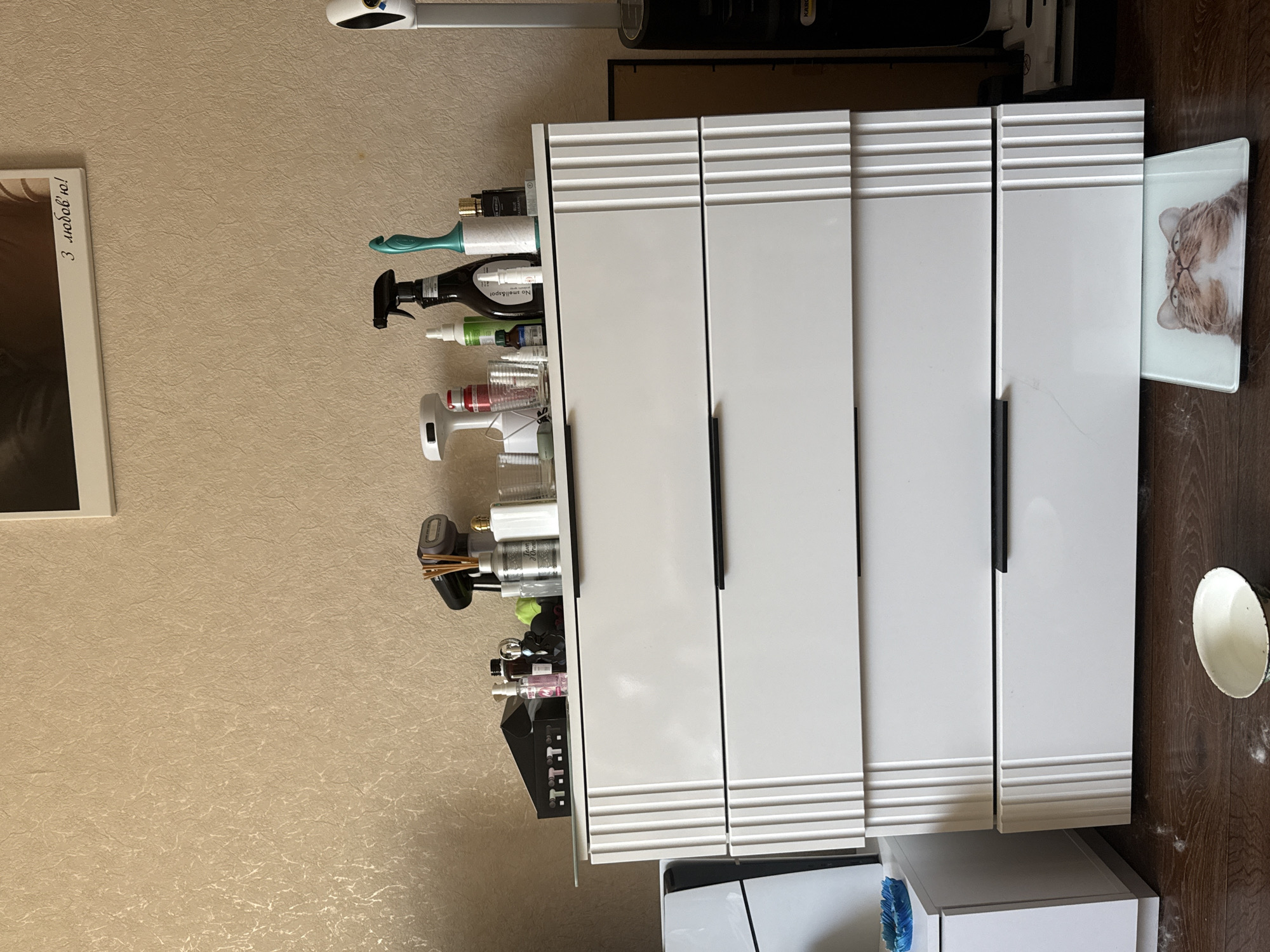 A dresser with four drawers stands in an indoor environment. The top of the dresser holds several items, including spray bottles, a hair dryer, and decorative objects. A framed picture hangs on the wall above the dresser. To the left of the dresser, a white cabinet is visible. The floor in front of the dresser is made of dark wood and is scattered with white debris. A small white bowl sits on the floor to the left of the dresser. A glass placemat with a cat's face printed on it is placed under the bottom drawer of the dresser. To the right of the dresser, a tall, white object is partially visible, and behind it, a black appliance can be seen. The room appears to be a bedroom or a living area.