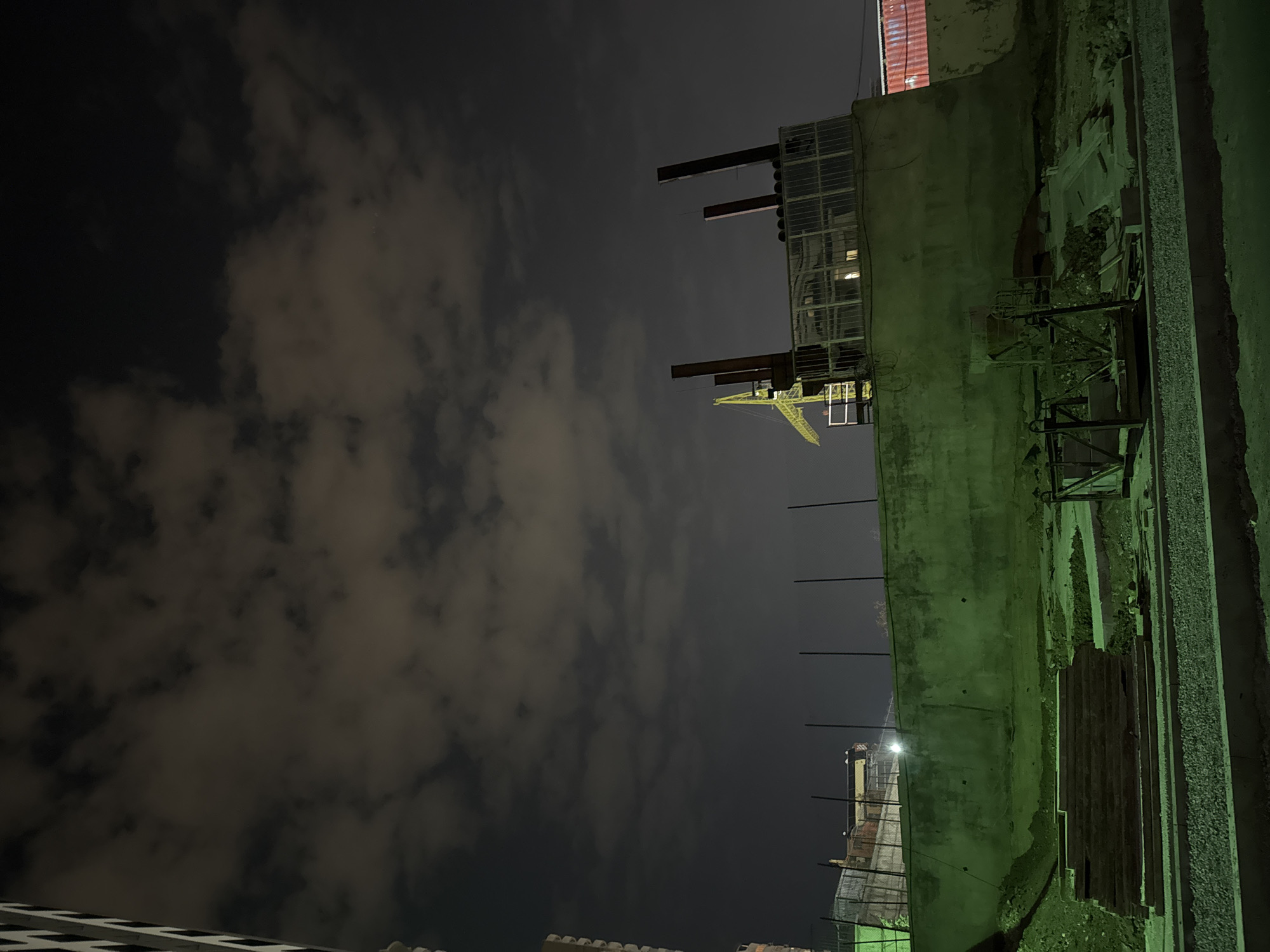 The image captures a nighttime scene, likely an urban industrial or construction area, presented in a portrait orientation. If rotated 90 degrees counter-clockwise, it reveals a landscape view looking upwards.
The upper left portion of the image (or top if rotated) shows a dark night sky with scattered, soft clouds that are faintly illuminated from below. Dominating the right side of the frame (or the bottom if rotated) is a long, textured concrete wall or building facade, rendered in a distinctive light-green color due to artificial lighting. Along the base of this wall, there is an accumulation of what appears to be dirt, debris, and stacks of wooden planks or other construction materials, along with some metal scaffolding or framework.
Behind the green wall, several structures rise into the dark sky. A yellow tower crane is visible in the mid-ground, its boom extending upwards. Several large, dark, vertical beams or pipes are prominently featured, extending from a building with a grid-patterned glass facade. Further in the background, dimly lit structures, including what looks like a red corrugated roof and distant city lights, can be observed. A bright, artificial light source is visible on the far left, behind a chain-link fence.
The scene is entirely illuminated by artificial light, contributing to the green cast on the foreground elements and creating a stark, quiet atmosphere. No people are visible, and no discernible activity is taking place. There is no visible text in the image. The location context is Yerevan, Armenia, suggesting this is a view of an urban construction or industrial zone there at night.