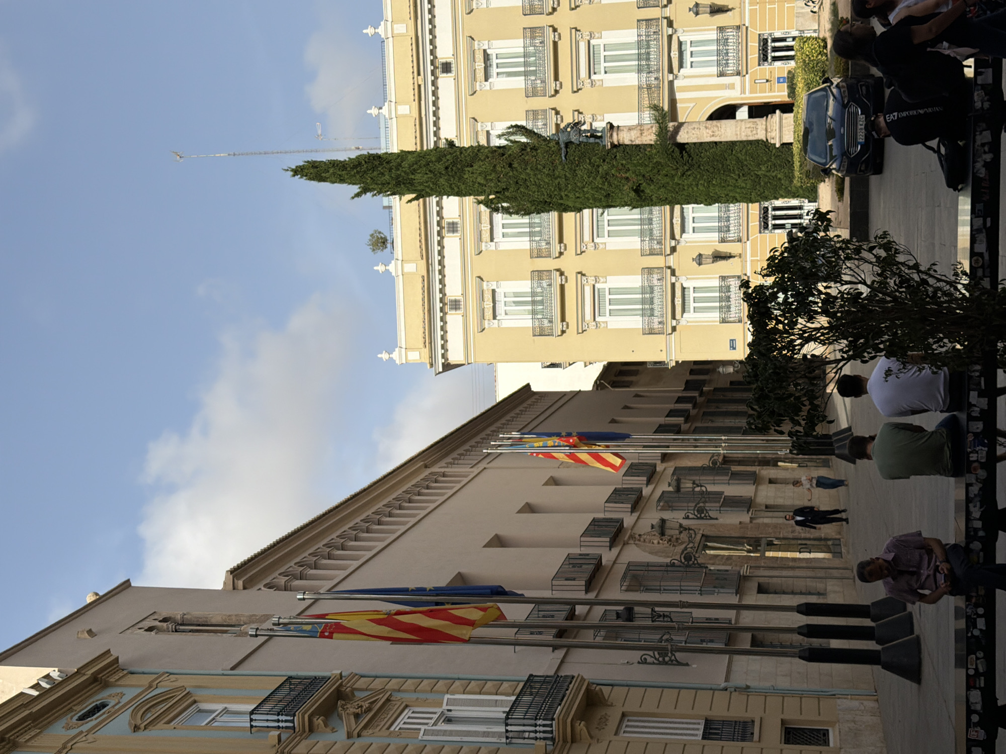 A sunny day shot captures a cityscape with ornate architecture and a tall cypress tree. In the foreground, people sit on a bench, some looking at their phones. The background features buildings with balconies and flags, and a dark-colored sedan is parked on the street.