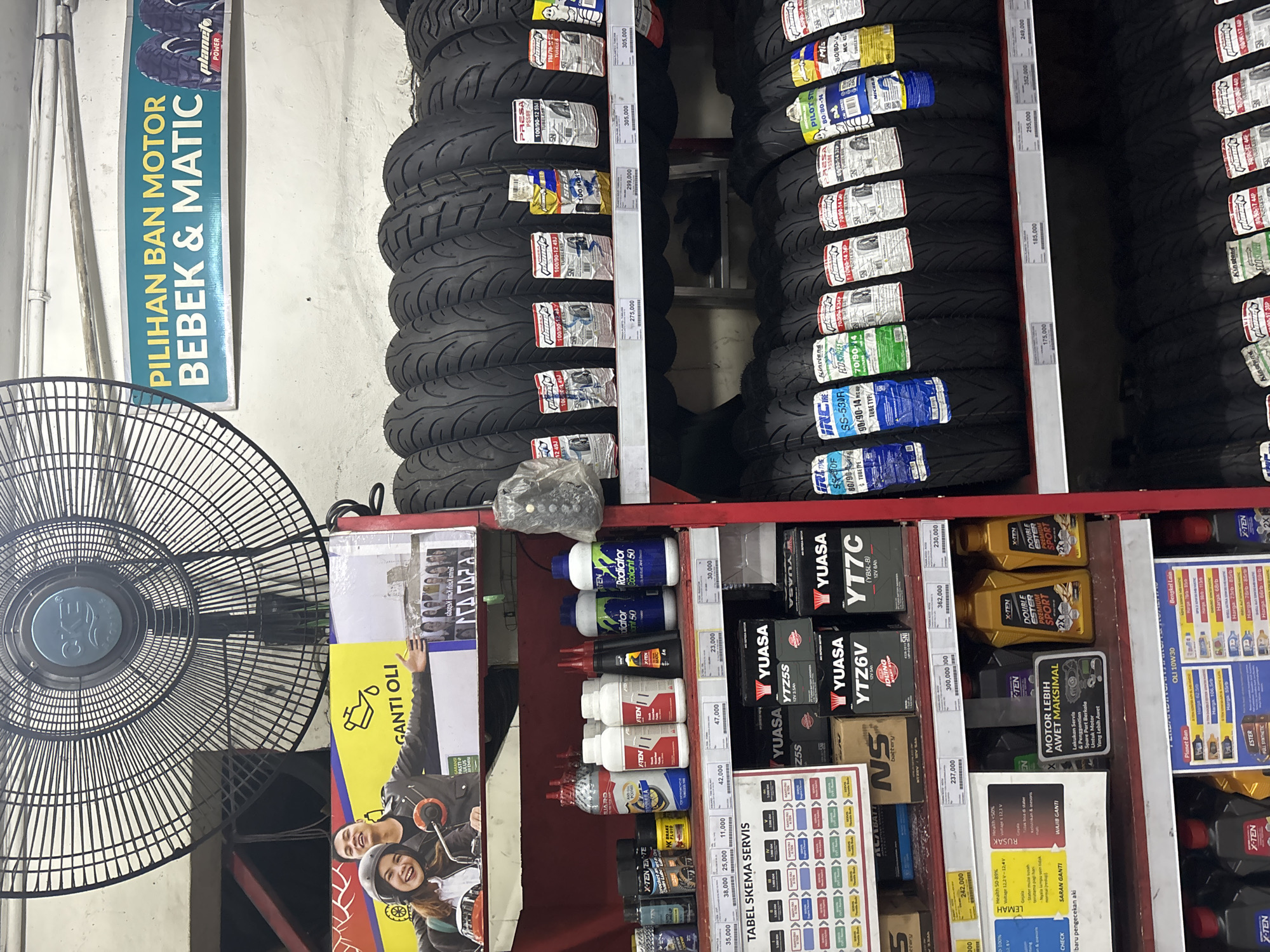 The image depicts the interior of a motorcycle parts shop, likely located in North Jakarta, Indonesia, given the context of common shop signage in the region. The scene is organized with shelves displaying various automotive products. In the foreground, shelves are stocked with motor oil bottles, coolant, and other fluid containers. A prominent sign displays "TABEL SKEMA SERVIS" (Service Schedule Table). Further back, rows of motorcycle tires are stacked, with some displayed with their packaging and pricing information. Above the shelves, a large industrial fan is mounted on the wall. A prominent banner with the text "PILIHAN BAN MOTOR BEBEK & MATIC" (Motorcycle Tire Choices for Automatic and Scooter Bikes) is visible. Another banner features an advertisement with images of young people on a motorcycle, promoting "GANTI OLI" (Change Oil). The overall environment suggests a retail setting focused on automotive maintenance and parts.