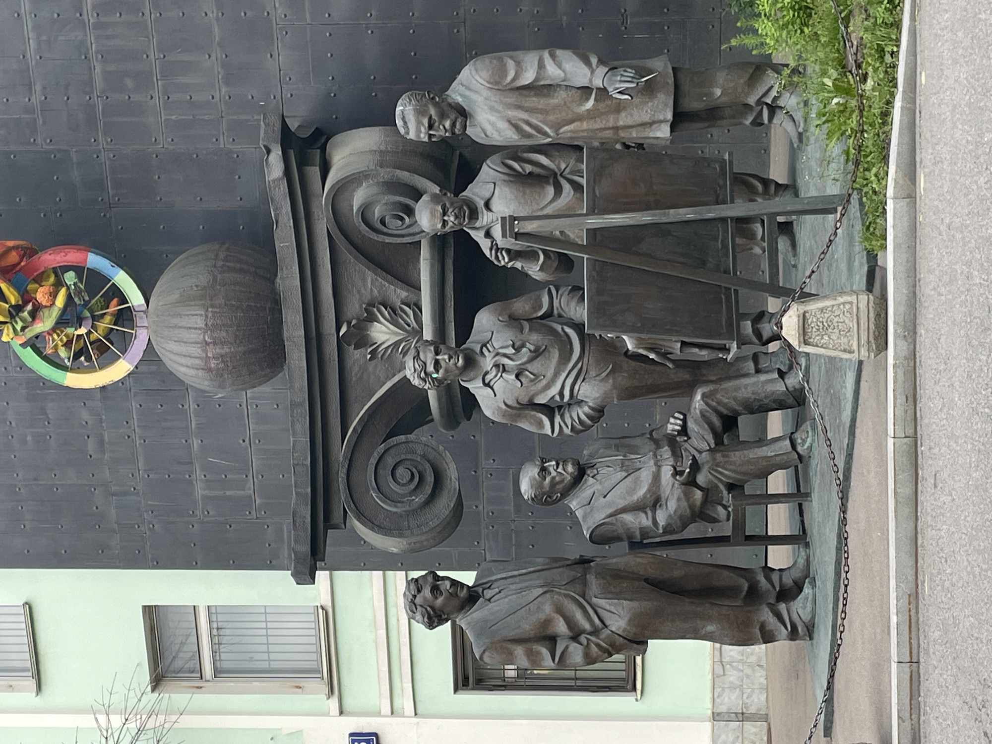 This is a full, eye-level shot of a bronze monument depicting a group of five men in what appears to be an art studio setting. One man is seated in a chair, facing right, while the other four stand around him. One of the standing men is in the process of painting on an easel. The monument is situated outdoors, with a light green building with windows to the left and a dark, textured wall with a large, ornate architectural detail behind it. A circular, colorful object is visible at the top of the dark wall. The monument is roped off with a chain on the left side. The scene appears to be located on a street or public square in Moscow, Russia, as indicated by a blue street sign with white lettering visible on the green building.