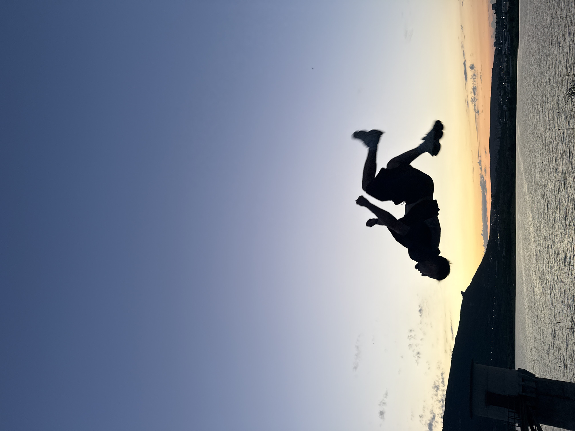 A silhouette of a person mid-air performing a flip is captured against a gradient sky at dusk. The sky transitions from a deep blue at the top to hues of orange and yellow near the horizon. Below the sky, a body of water reflects the fading light. In the background, a silhouetted landscape features rolling hills and a distant cityscape. To the left, a prominent concrete structure, likely part of a water tower or pumping station, stands near the water's edge. The person is in athletic attire, with arms and legs extended as they rotate. The overall scene suggests an outdoor recreational activity taking place near a body of water with urban and natural elements in the backdrop. The specific location in Shumen, Bulgaria, is not visually identifiable from the image itself.
