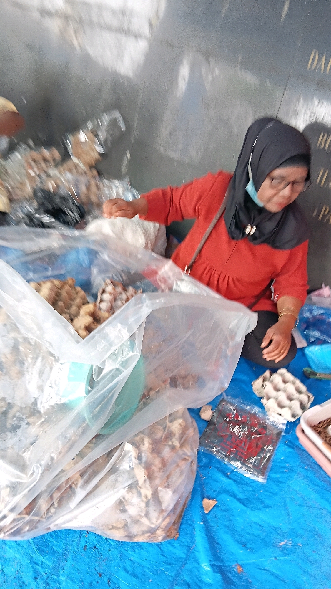 A woman, appearing to be of middle age, is seated on a blue tarp, likely outdoors, in what could be a market or roadside stall setting in Batusangkar, Indonesia. She is wearing a red long-sleeved top, a black hijab, glasses, and a light blue face mask. Her left hand is reaching into a large clear plastic bag filled with dried goods, possibly food items like ginger or herbs. Another large clear plastic bag, also containing similar dried goods, is positioned in front of her. To her right, on the blue tarp, are several items: an egg carton containing white eggs, a smaller plastic bag with red lettering, and a pink plastic container. Behind her, a dark wall, possibly a building or fence, is visible, with some indistinct writing or markings. The overall scene suggests a commercial transaction or preparation for sale of produce.