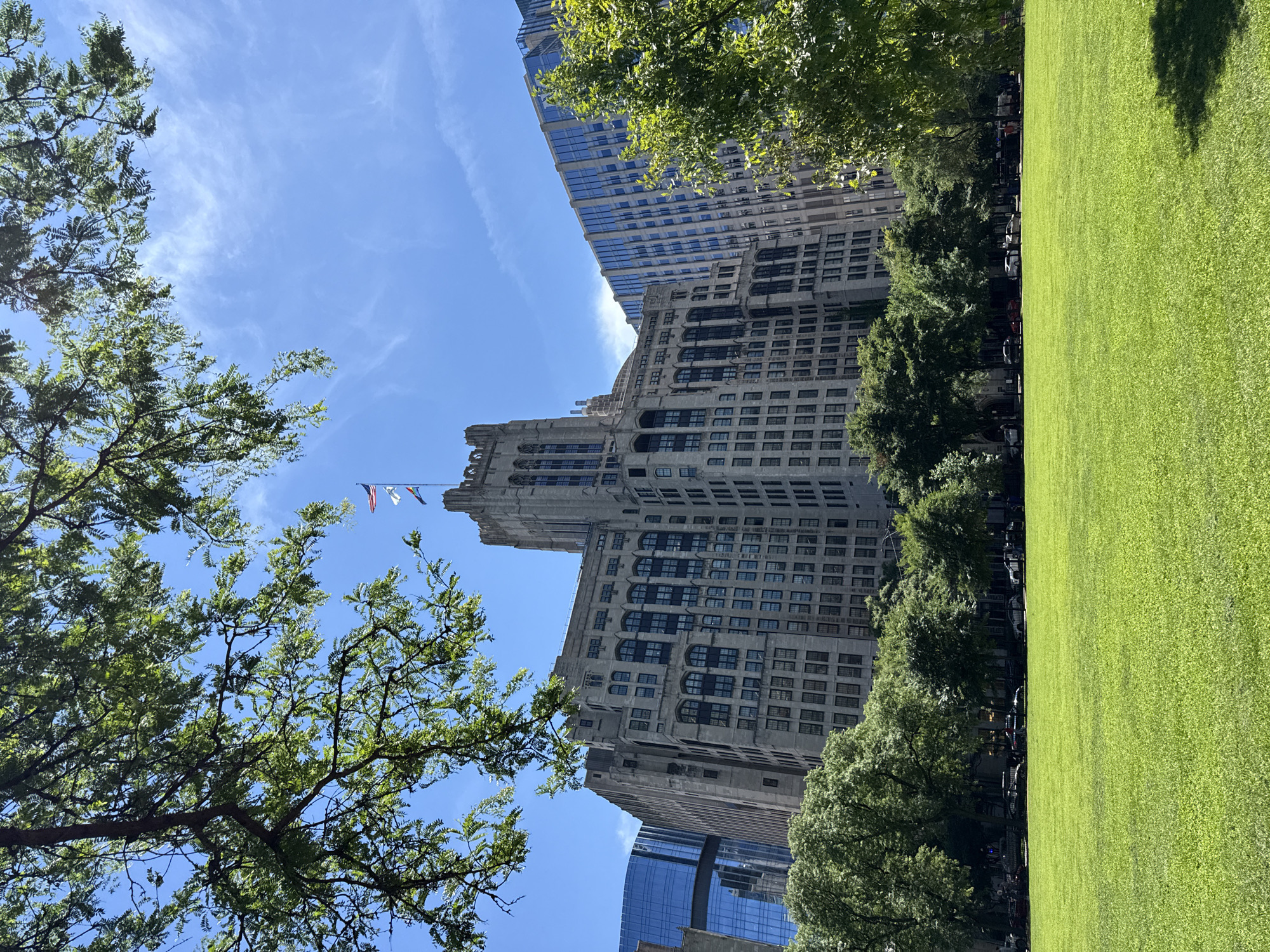 This image captures a sunlit day in a cityscape, likely Chicago, United States of America, given the architectural style of the buildings and the presence of a manicured lawn. The scene is a juxtaposition of nature and urban development.
Dominating the background are tall, multi-story buildings with distinct architectural features. One prominent building appears to be made of stone or concrete, with numerous windows and a Gothic-inspired tower. Another, more modern building, features a facade of reflective blue glass. Trees with lush green foliage are scattered throughout the scene, some framing the buildings and others lining a pathway.
To the right of the image is a large, vibrant green lawn, bathed in sunlight. The bright green of the grass suggests it is well-maintained and possibly part of a park or public square. The sunlight casts subtle shadows from the trees onto the lawn, indicating it is daytime, likely late morning or early afternoon. The sky above is a clear blue with wispy white clouds.
In the distance, several flags are visible, fluttering against the sky. These include what appears to be the American flag and possibly a state or city flag. The flags suggest a public space where civic symbols are displayed.
While no distinct individuals are clearly discernible, there are hints of people or activity beneath the trees and along the pathway adjacent to the lawn, with some parked cars visible further back. The overall mood of the image is serene and peaceful, highlighting the blend of urban greenery and impressive architecture. The bright sunshine and clear sky contribute to a positive and inviting atmosphere.