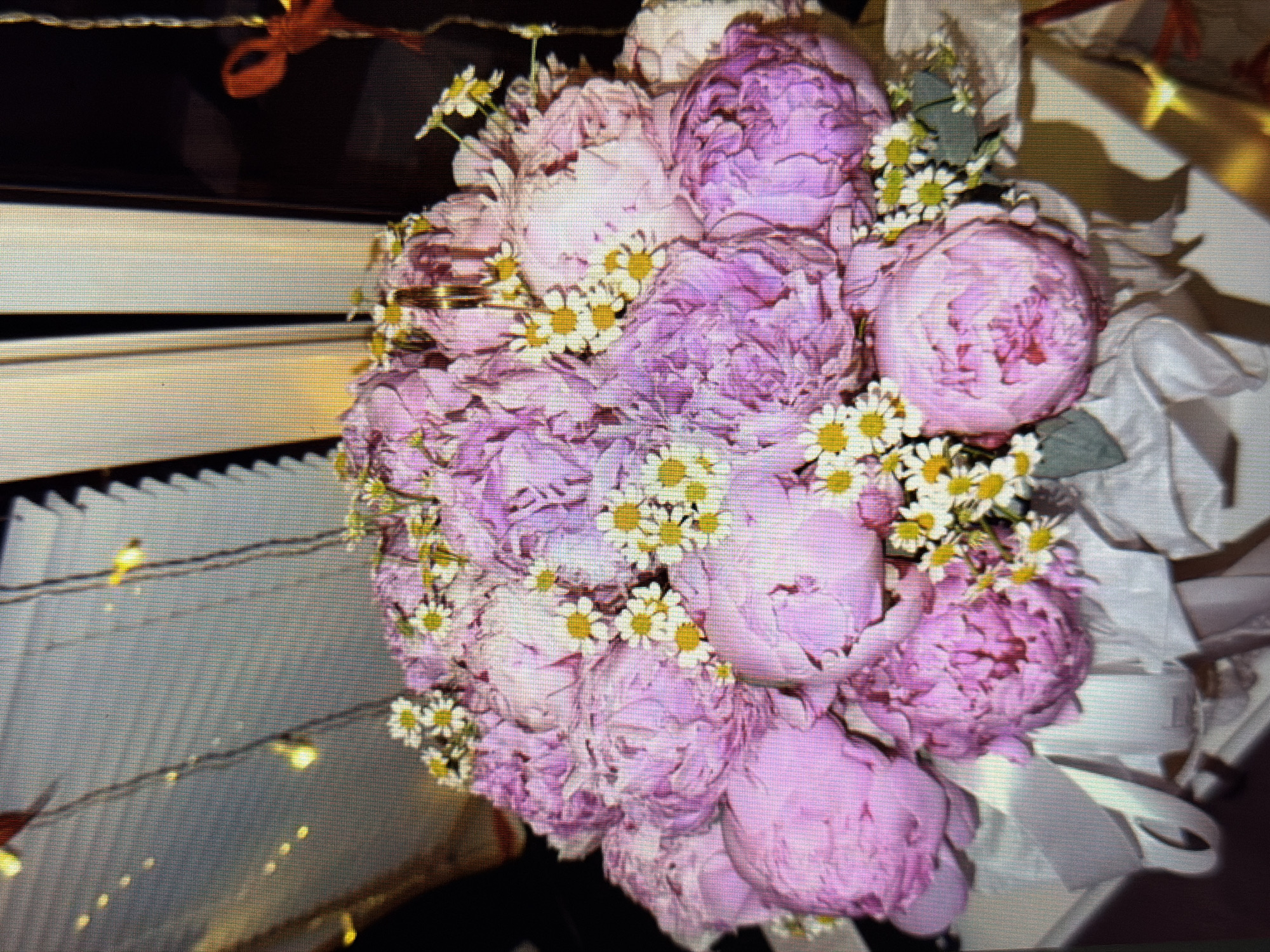 The image shows a close-up of a bouquet of pink peonies and small white daisies, arranged in a white paper wrapping. The bouquet is likely indoors, with soft lighting and a blurred background that suggests a window or decorated wall. There are no people or specific landmarks visible. The scene is static and does not depict any action or interaction. The location in the city of Sirius, Russia, is not discernible from the image.