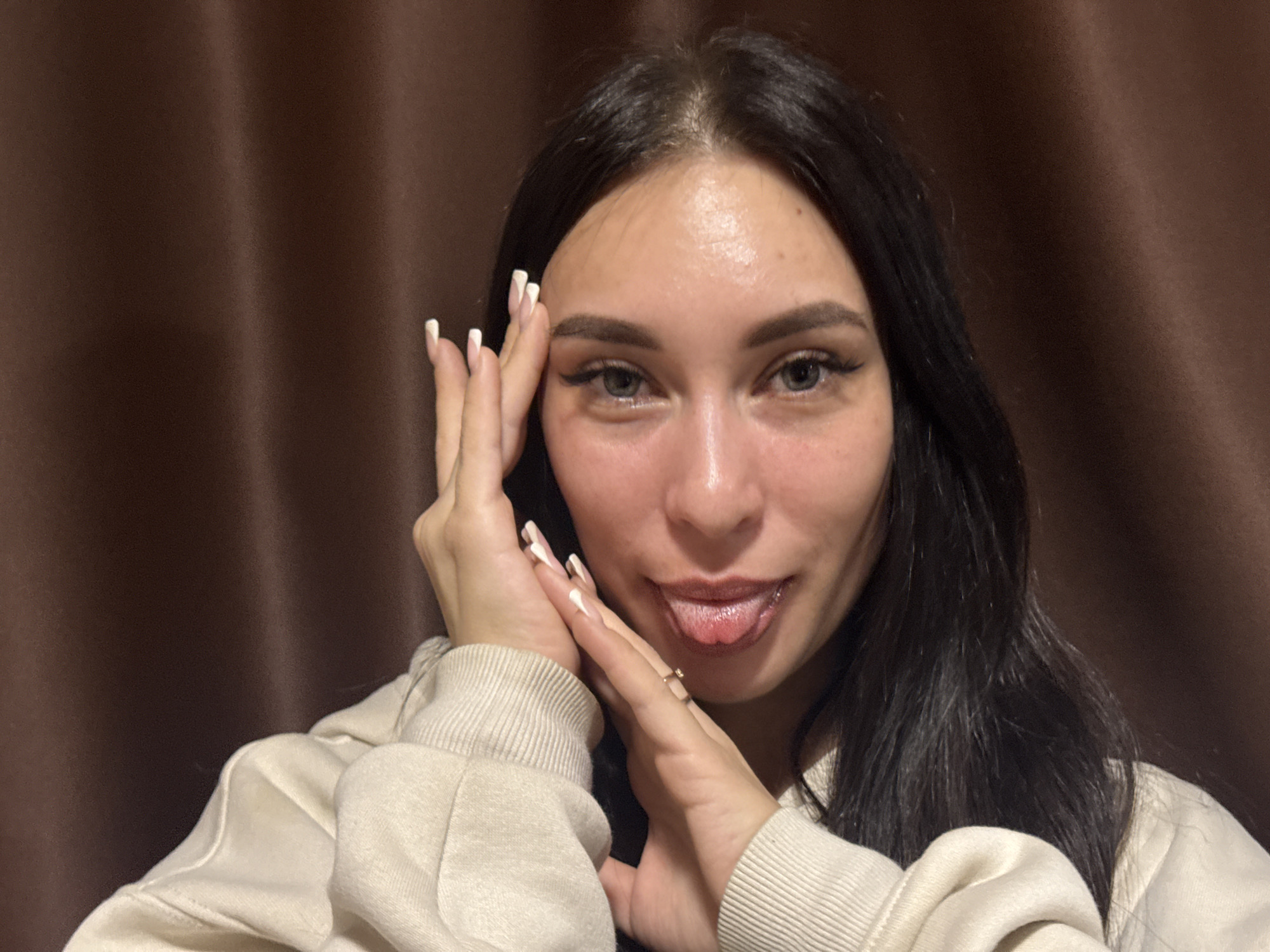 A young adult woman is centered in the frame with a brown draped background. She has fair skin, dark hair, and her tongue is slightly protruding from her mouth. Her hands are held up near her face, with manicured fingernails painted in a white and neutral design. She is wearing a light-colored sweatshirt. The location in Chaikovsky, Russia, cannot be determined from the image.