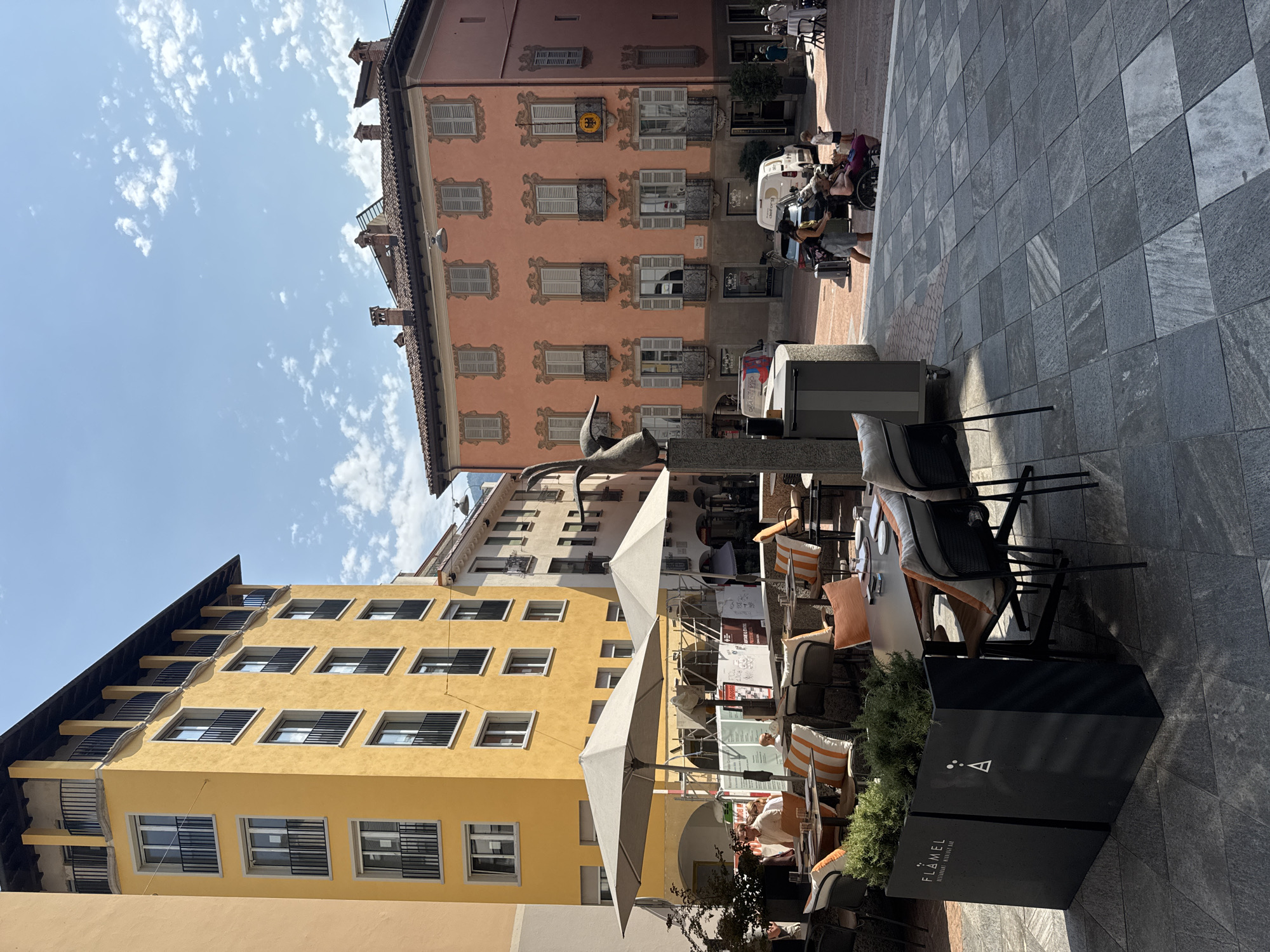 The image captures a sunny day in a city square, likely in Lugano, Switzerland. A brightly colored yellow building with balconies and numerous windows dominates the left side of the frame, with a pale orange building of similar architectural style on the right, featuring decorative window frames and shutters. In the foreground, an outdoor dining area is set up with tables and chairs, some adorned with orange and white striped cushions. Large umbrellas provide shade over some of the tables. A dark planter box in the lower left corner displays the text "FLAMEL RESTAURANT". People are visible in the background, some walking and others seated at tables. A woman is pushing a person in a wheelchair, and a young child stands nearby. Several vehicles, including cars and a white van, are parked or moving in the distance. The ground is paved with large, square tiles. A sculpture of a bird-like figure stands to the left of the orange building. The sky is blue with scattered white clouds. The scene suggests a vibrant and pleasant urban environment.