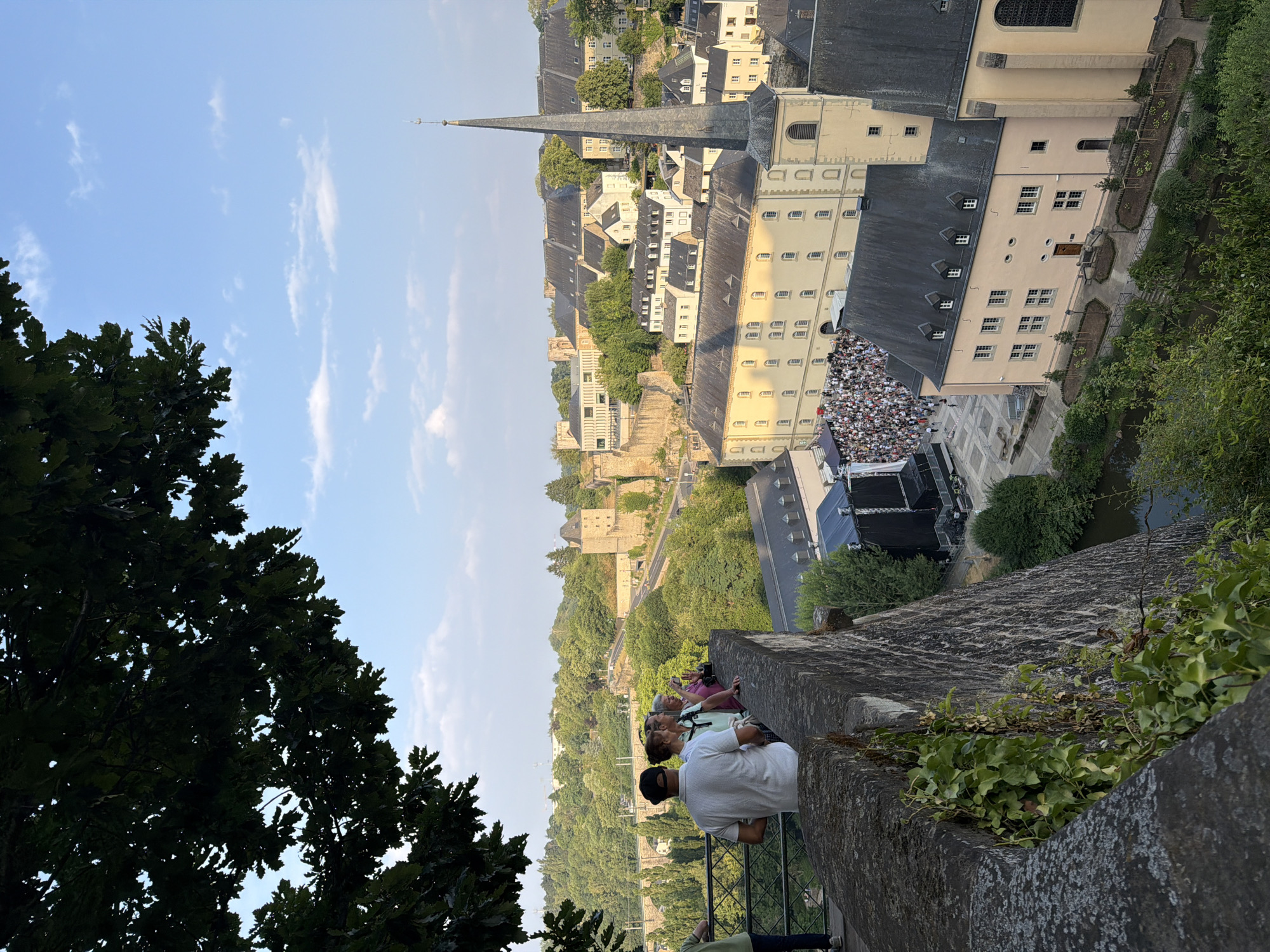 An outdoor, elevated shot captures a crowd gathered in a square below, adjacent to a large building with a steeple. A group of people are visible on a stone wall in the foreground, looking out over the scene. The city of Luxembourg is recognizable by its distinctive architecture, including historical buildings and fortifications. The time of day appears to be late afternoon or early evening, with the sun casting warm light on the buildings. Green trees and foliage surround the urban landscape, adding a natural element to the scene. The sky above is a clear blue with a few scattered white clouds.