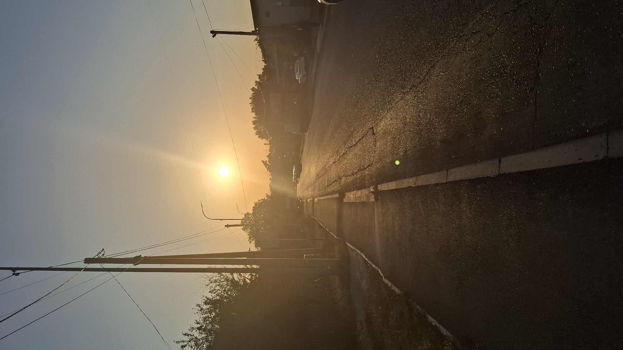 The image depicts a street scene at sunset in Shymkent, Kazakhstan. The sun is low on the horizon, casting a warm, golden light that illuminates the asphalt road and a sidewalk. Power lines stretch across the clear sky, supported by utility poles. Trees line the left side of the road, and buildings are visible in the distance on the right. Several cars are parked or driving on the road. The overall impression is one of a quiet, late afternoon in a residential or semi-urban area.