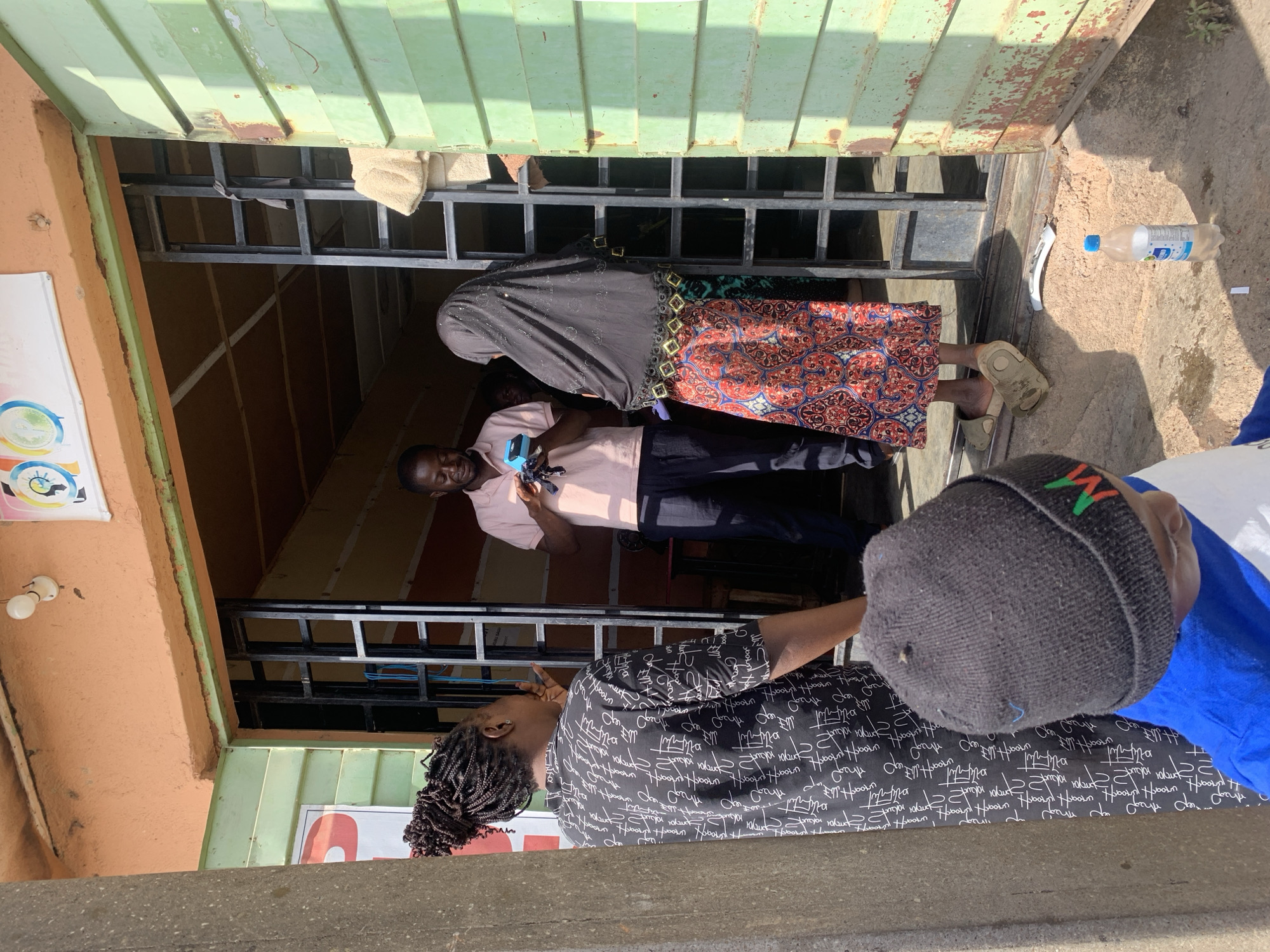 The image depicts several individuals in what appears to be an entry to a building in Jos, Nigeria. A man is visible inside the entrance wearing a pink shirt and dark pants, holding a blue object in his hand. A woman wearing a black headscarf and a patterned skirt with a border of gold squares is also entering. Another woman with braided hair, wearing a black and white patterned shirt is standing outside the entrance. Beside her, another individual, only partially in frame, is wearing a gray beanie. A plastic water bottle is on the ground outside the entrance. The building has a light green exterior.