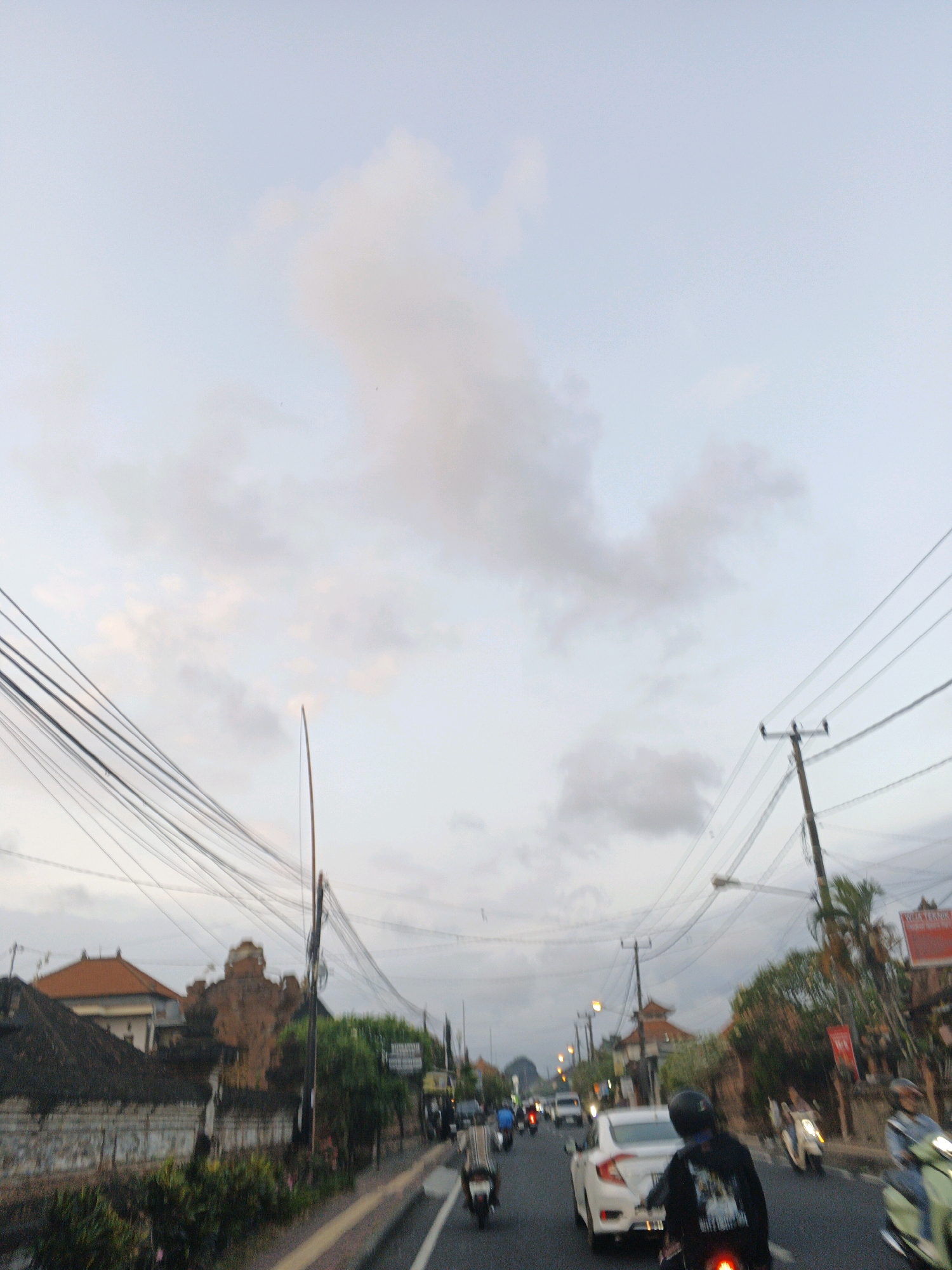 The image depicts a street scene in Denpasar, Indonesia, likely during the late afternoon or early evening, given the soft lighting and overcast sky. The perspective is from a moving vehicle, possibly a motorbike, looking forward along a road bordered by buildings and dense utility poles with numerous wires.
On the left side of the road, there are traditional Balinese-style buildings with terracotta roofs and stone walls. Lush green foliage and vegetation are visible along the roadside. On the right side, more buildings are present, including a shop with a red sign. Palm trees and other tropical vegetation add to the landscape.
The road itself is moderately busy with traffic. Several motorbikes are in motion, some carrying single riders and others with passengers. A white car is also visible, as is a light-colored scooter with a rider. People are wearing helmets and casual clothing. The overall impression is of a typical urban street with a vibrant flow of daily life. The environment appears warm and humid, characteristic of a tropical climate.