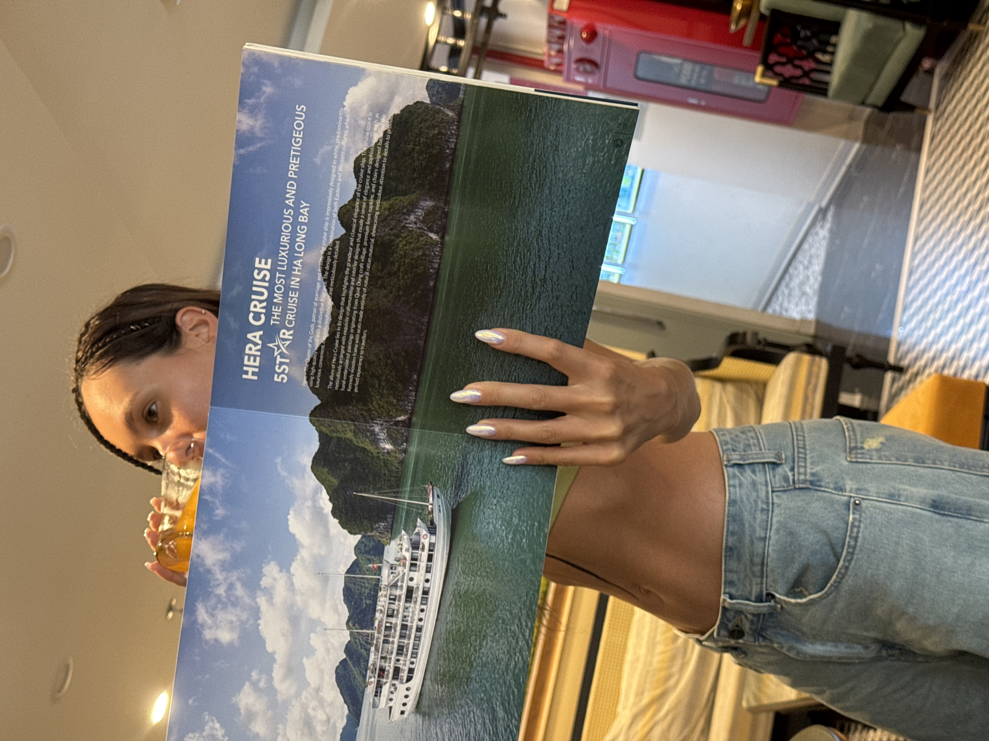 A woman with braided hair drinks from a glass while reading a magazine titled "Hera Cruise," which advertises a 5-star cruise in Ha Long Bay. The advertisement includes a photo of the cruise ship and Ha Long Bay. The woman is wearing light blue jeans and a green top. The background includes a red oven and a patterned chair. The location is likely Hanoi, Vietnam, based on the cruise destination being in Ha Long Bay.