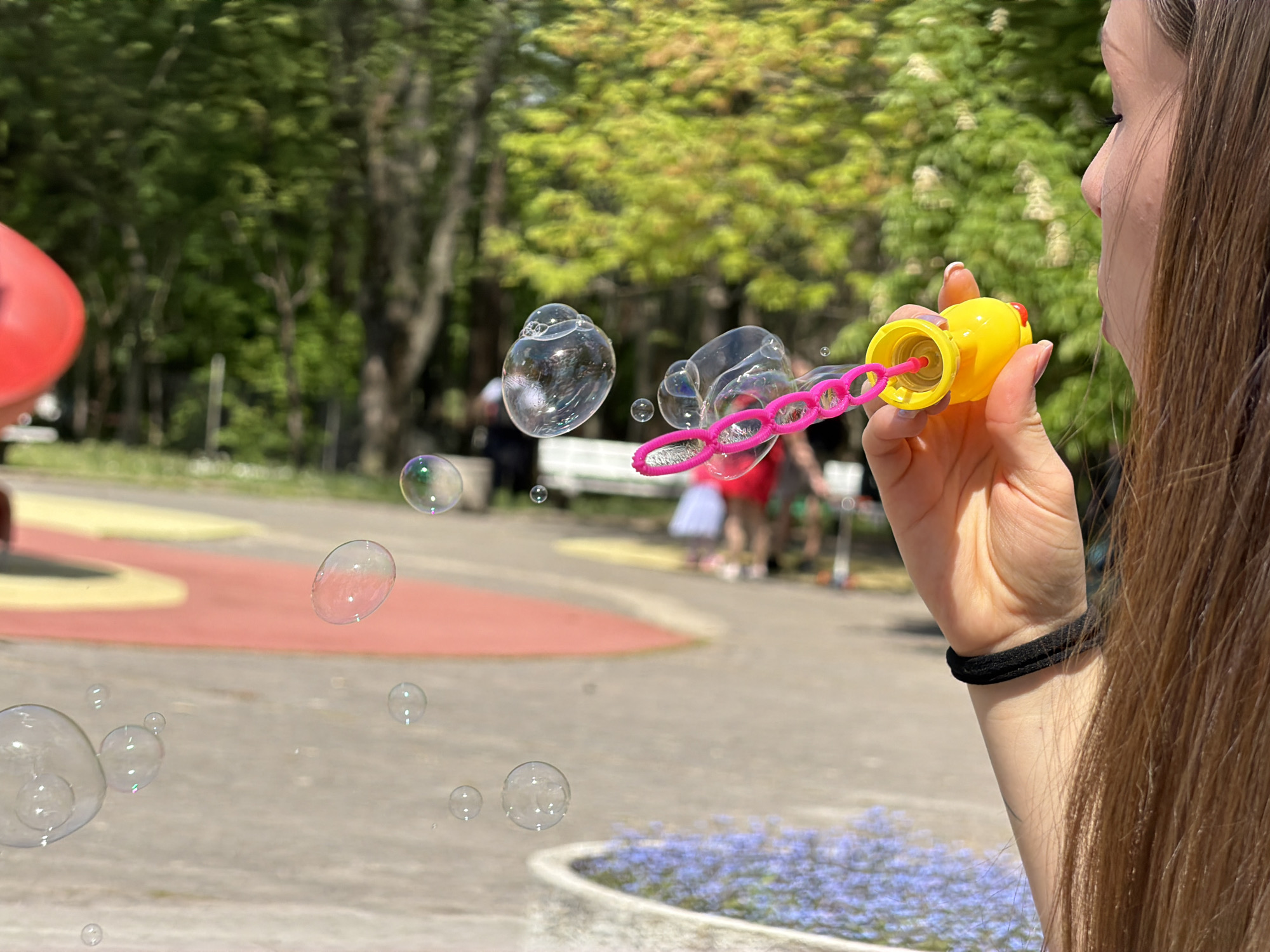 A woman is blowing bubbles in a park. She is holding a yellow bubble wand and has just blown a cluster of bubbles. More bubbles are floating in the air. In the background, there is a playground with a red structure, a white bench, and other people. The location appears to be a park in Sofia, Bulgaria. The environment is sunny, with green trees and blue flowers planted in a circular pattern near the bottom of the frame. The woman is wearing a black hair tie on her wrist and her long brown hair hangs down the right side of the image.