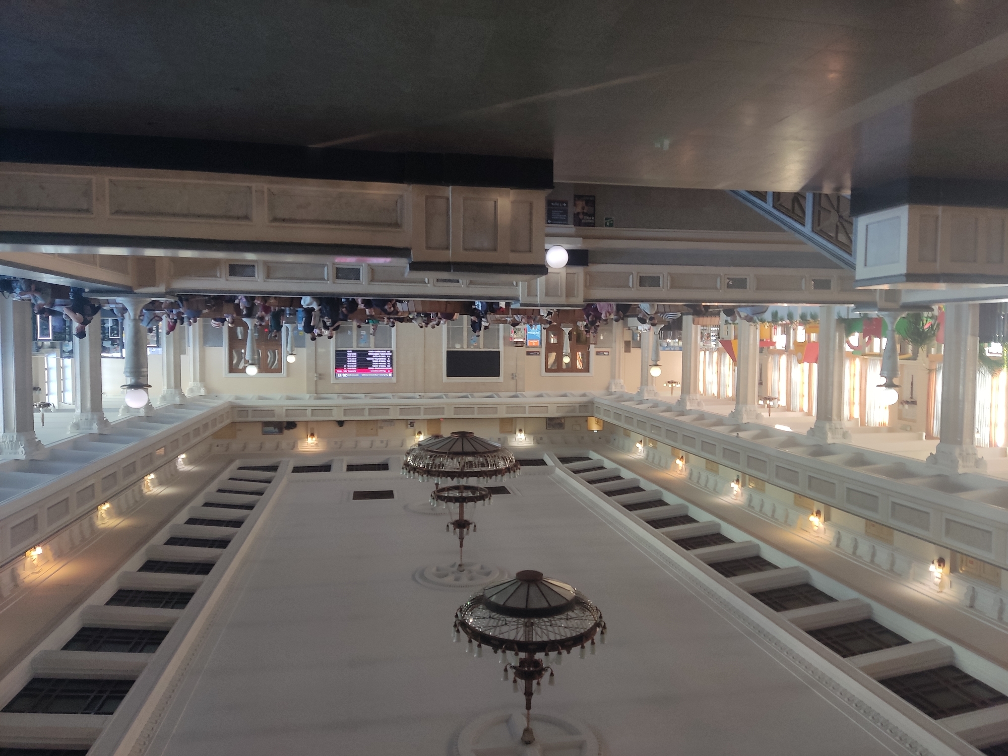 The media file depicts an interior shot of a grand, multi-story building, likely a public space such as a train station or a museum, given the architectural style and the presence of people. The image is rotated 90 degrees counterclockwise.
The scene is characterized by its high ceilings, white ornate columns, and decorative railings along the upper levels. Ornate chandeliers with glass shades hang from the ceiling, illuminated by warm lighting. The upper floors feature rows of windows and architectural details. A prominent feature is the wide, white-railed balcony that encircles the central open space. Below, on the ground level, are visible signs of activity, with people milling about. Some are seated, possibly waiting, while others appear to be walking. A digital display board with red text is visible on the left side of the frame, suggesting a transportation hub.
The lighting suggests it is daytime, but the interior setting makes it difficult to determine the weather. The overall impression is one of a well-maintained, classic public building. The mood seems neutral, with no strong emotional cues conveyed by the subjects.
Visible text includes "30:13" on the digital display board. The context provided indicates the location is Novosibirsk, Russia, which aligns with the architectural style often found in historical Russian public buildings.