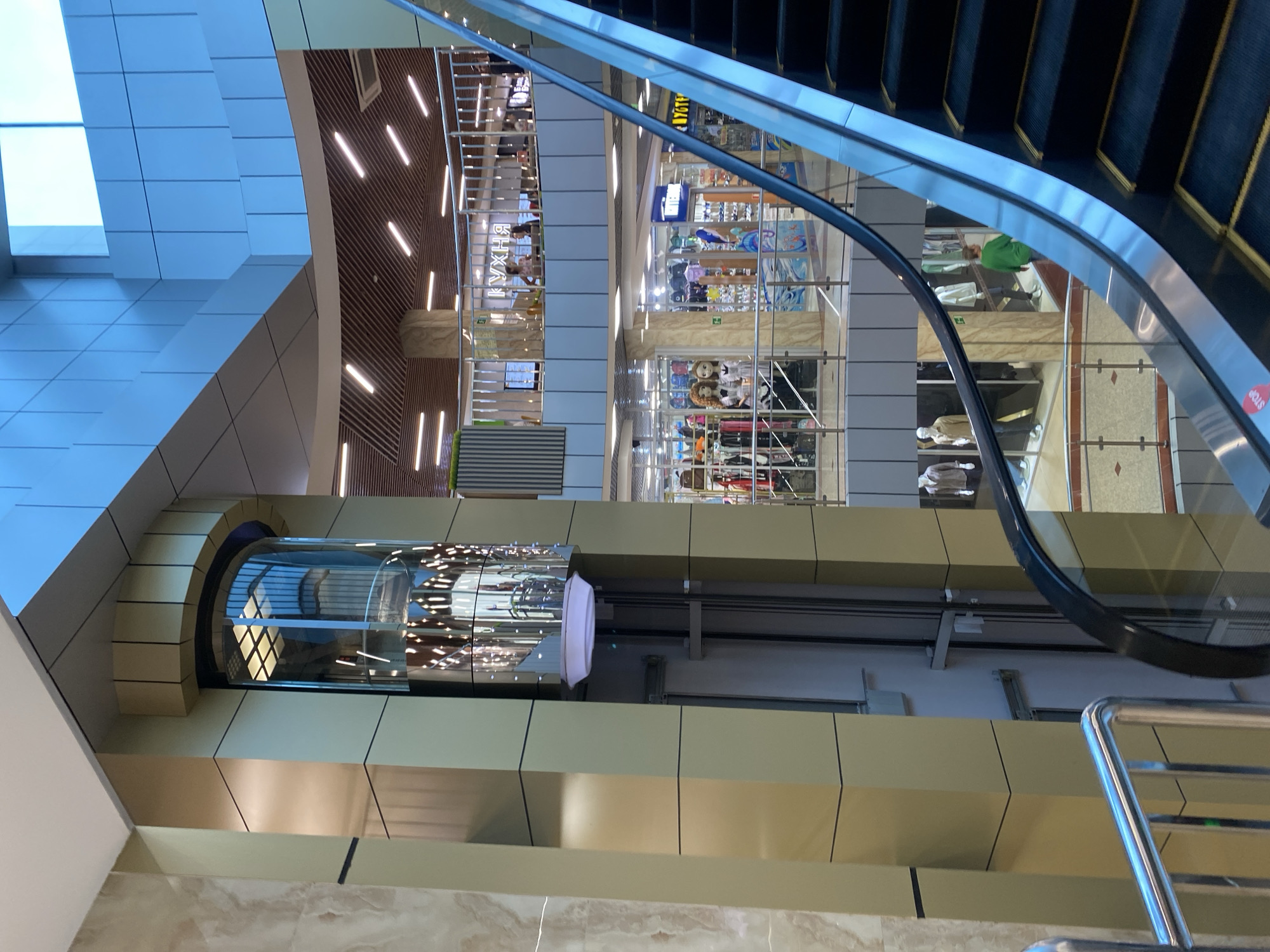 The image shows a modern indoor space, likely a shopping mall, with multiple levels and escalators. A glass elevator is visible on the left, encased in a golden-colored pillar. Escalators run diagonally across the frame. Several shops are visible on the lower levels, displaying mannequins and merchandise. People are seen on the upper levels, some seated at tables. The architecture features clean lines, blue and beige color schemes, and ample lighting. The overall impression is one of a bustling commercial environment.